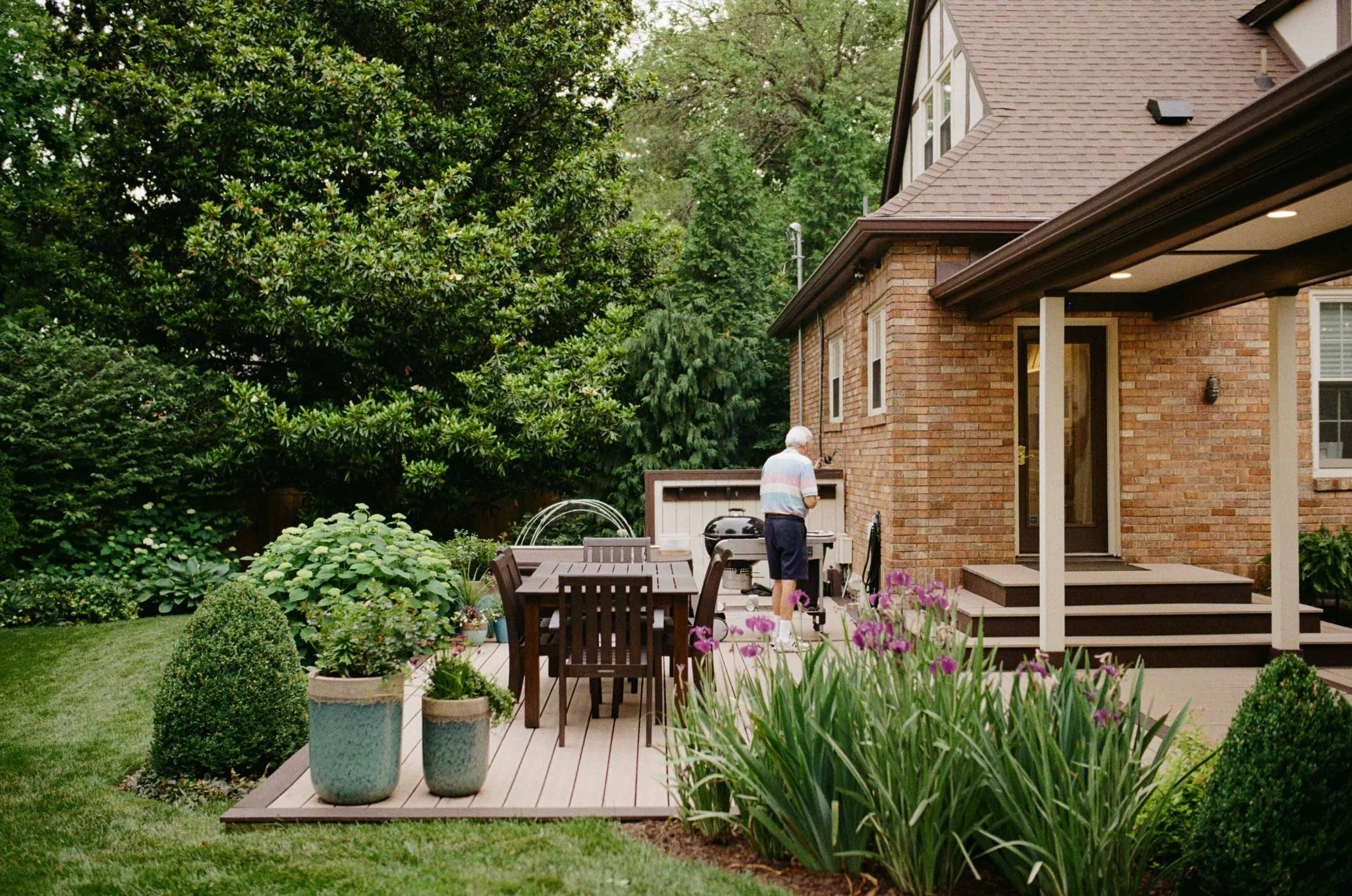 A man in a striped shirt and shorts grilling in his backyard, surrounded by potted plants and lush green trees.