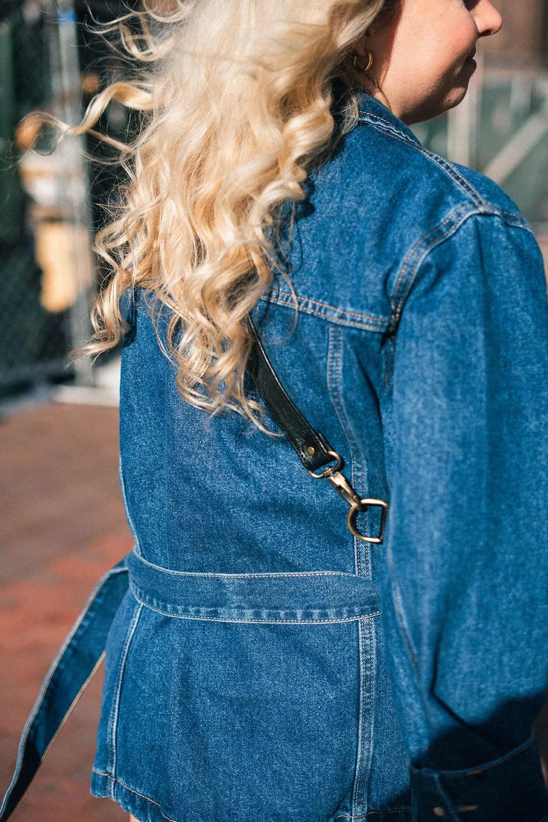 Editorial style photo of a close-up of a woman with blonde, curly hair wearing a blue denim jacket in the bright sun.