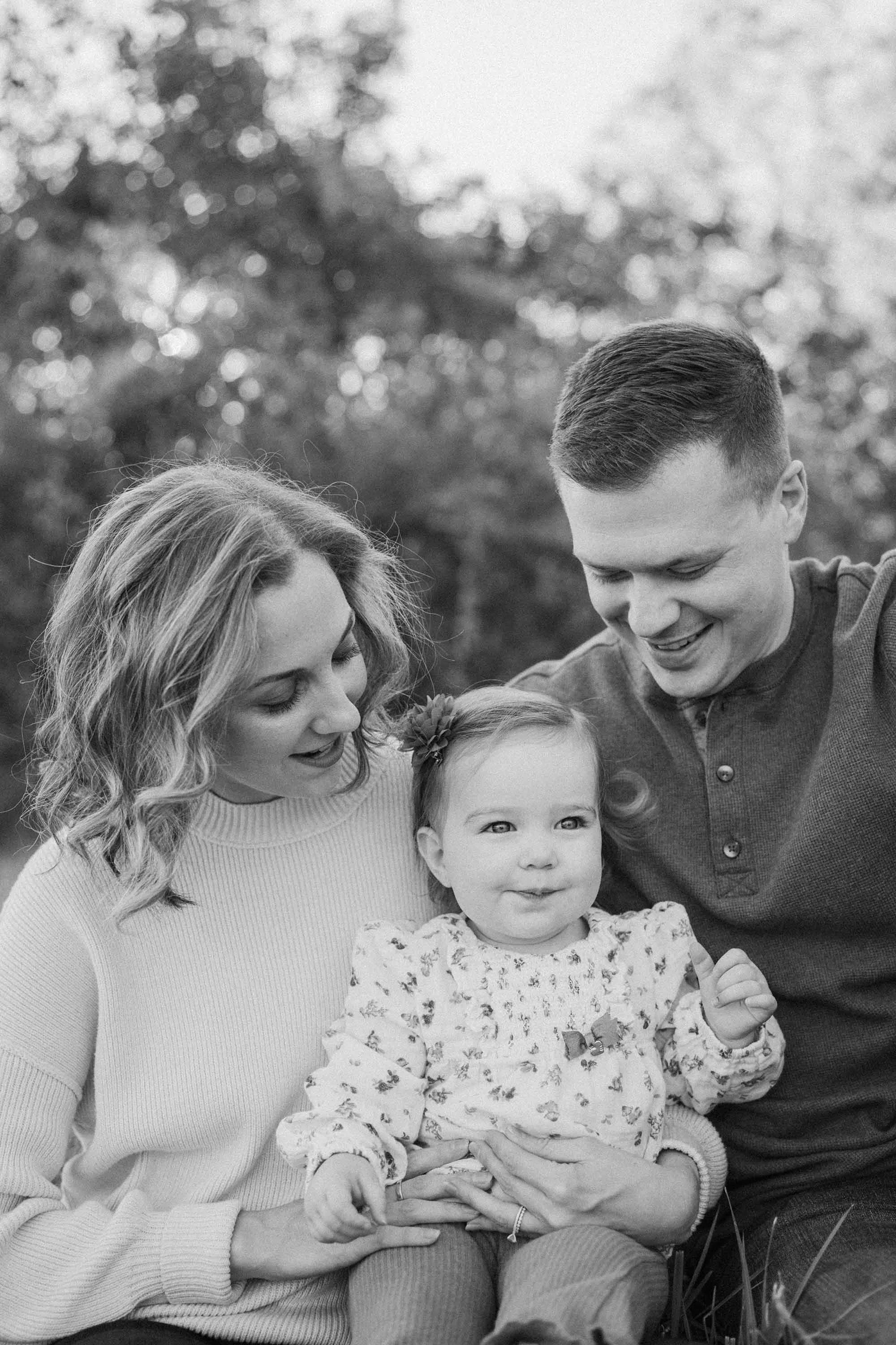 A black and white photo of a family of three sitting outdoors, consisting of a mother, a father, and a young daughter. All are smiling and looking at each other with a blurred natural background.