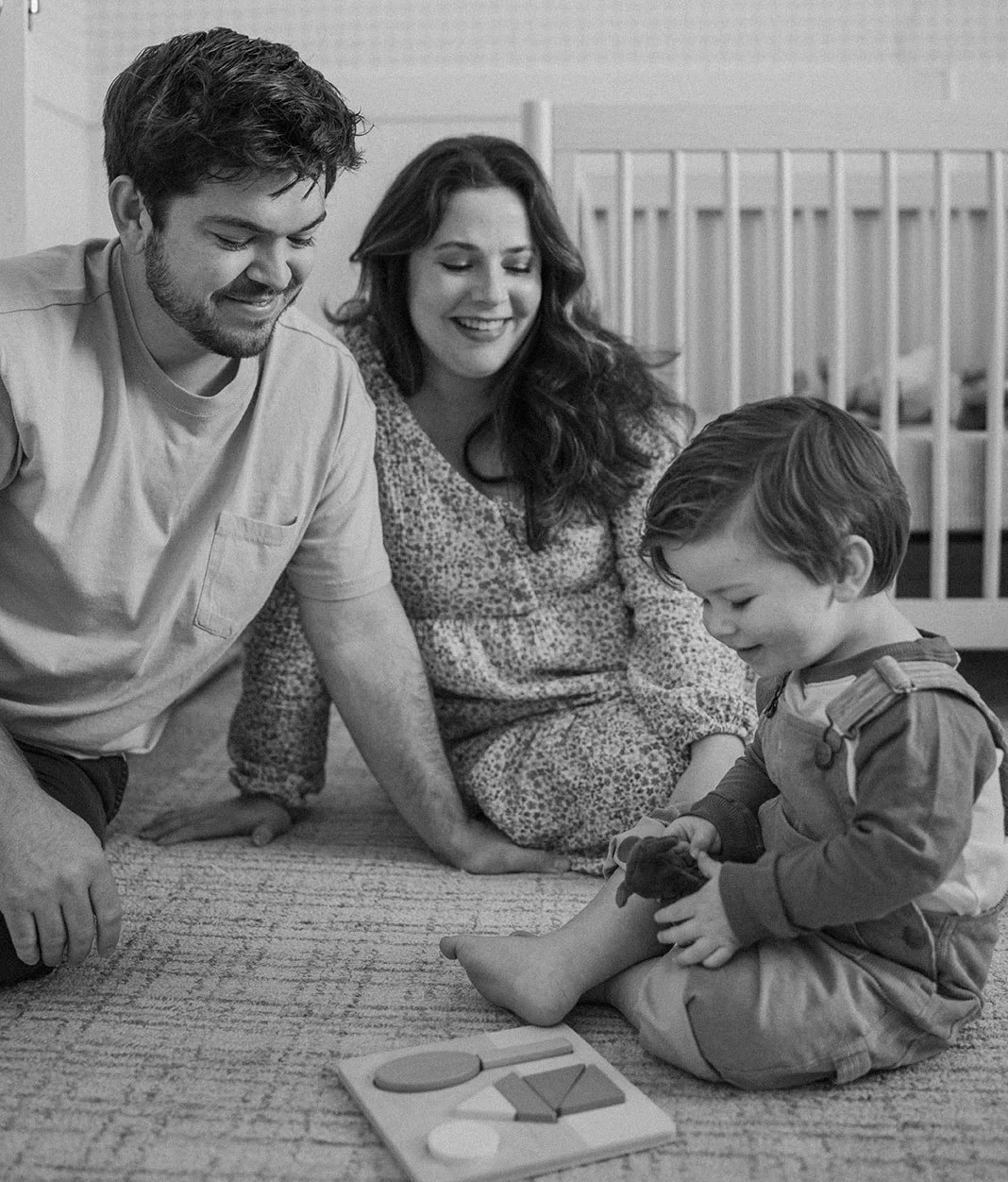 A family of three, including a smiling woman, a smiling man, and a young boy, playing with a toy puzzle on the floor in a nursery.