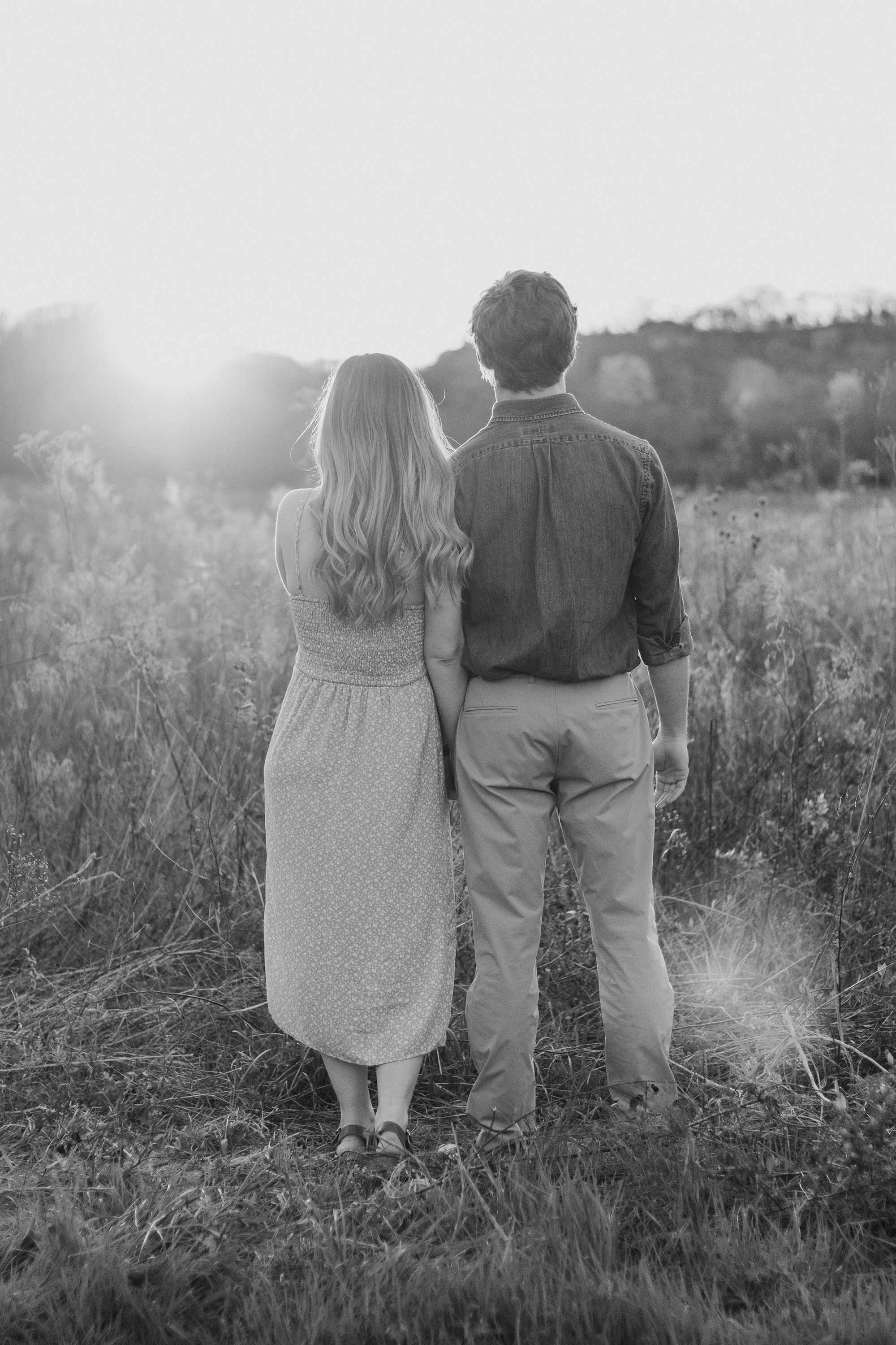 A black-and-white photo of a couple standing in a field, viewed from behind, holding hands, with the sun setting in the background.
