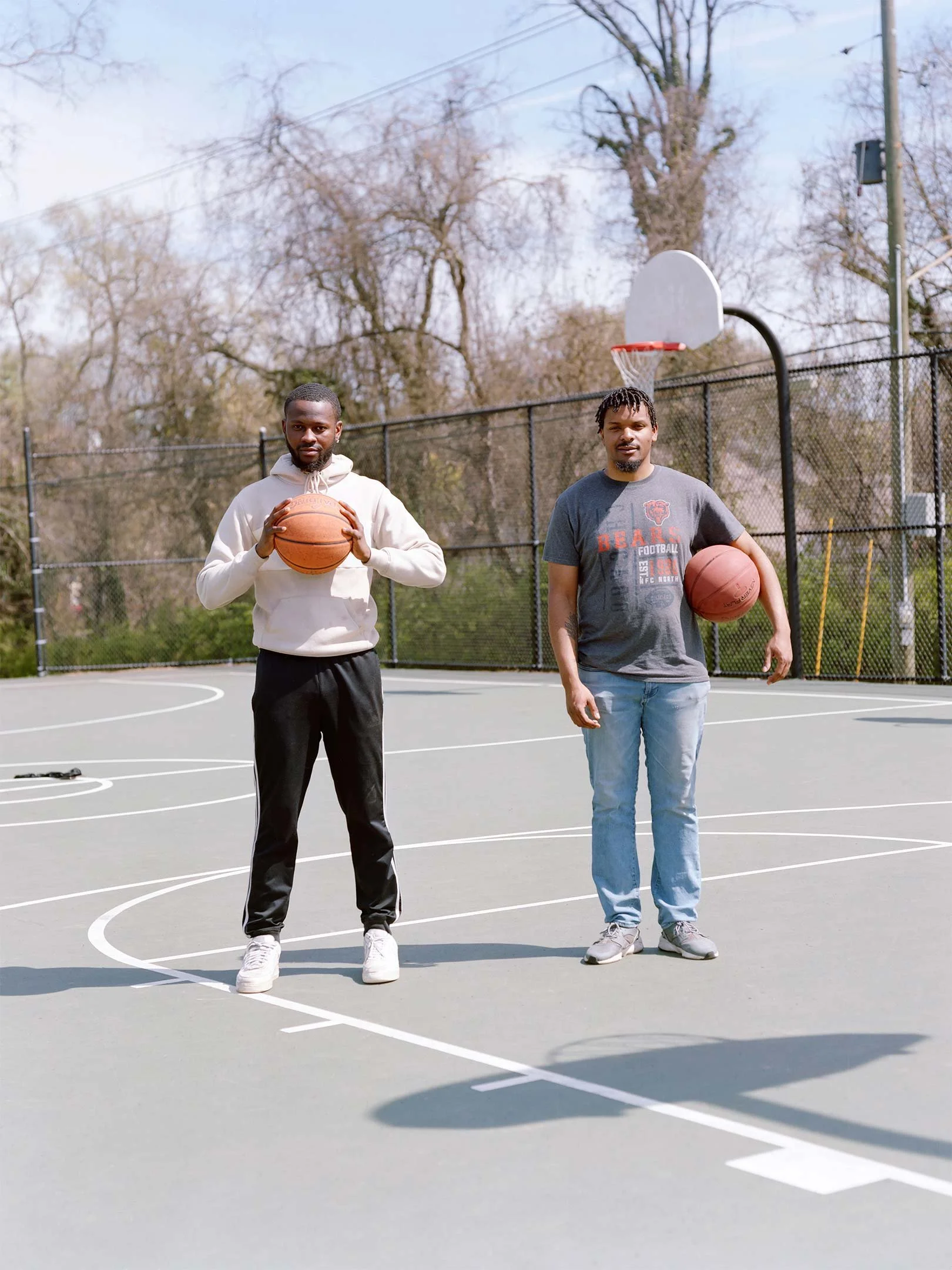 Two men standing on an outdoor basketball court holding basketballs, with a basketball hoop and a chain-link fence in the background.