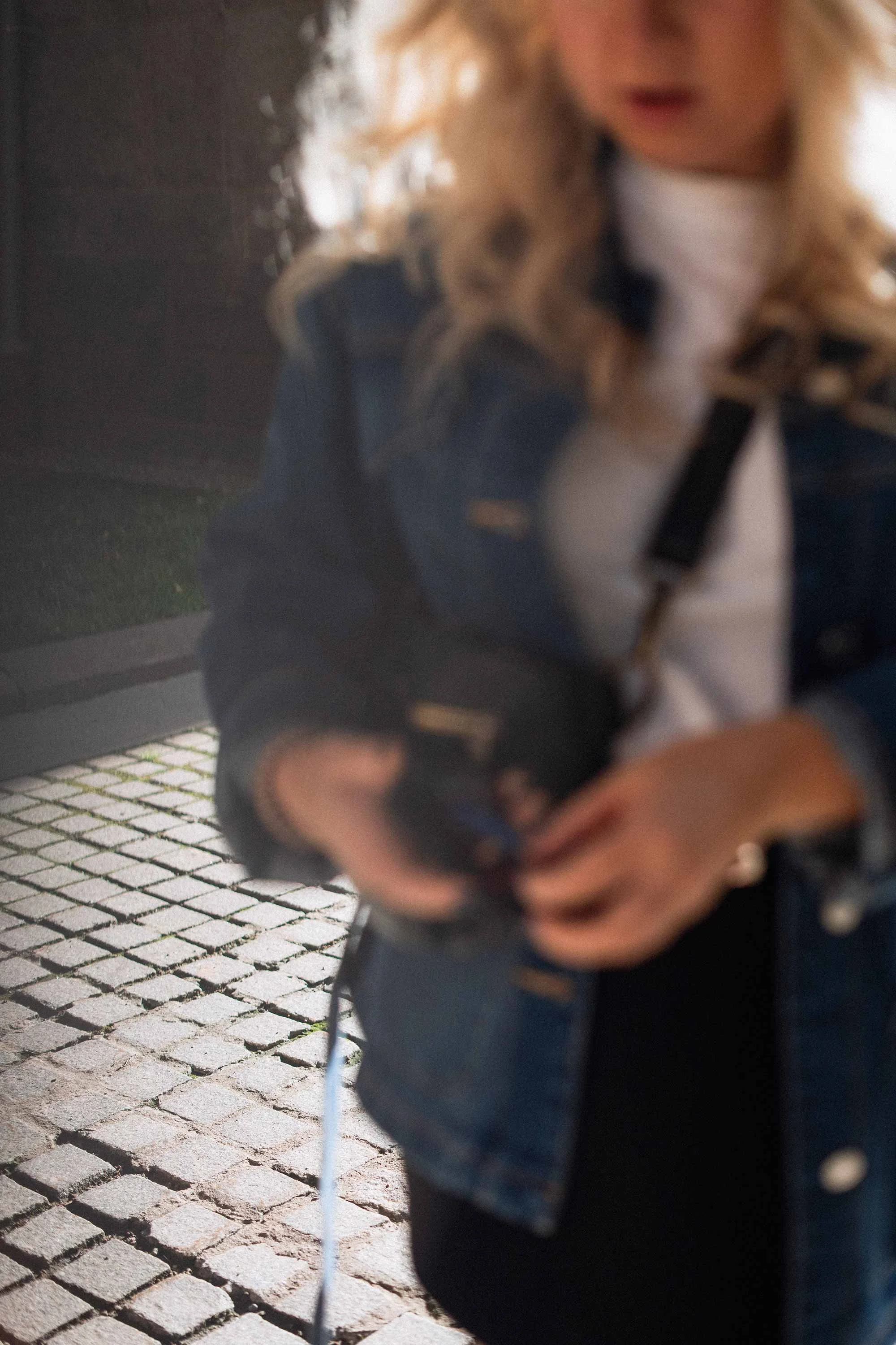An editorial style, blurred photo of a young woman standing outdoors in the sun, in a denim jacket with curly blond hair putting something in her purse.