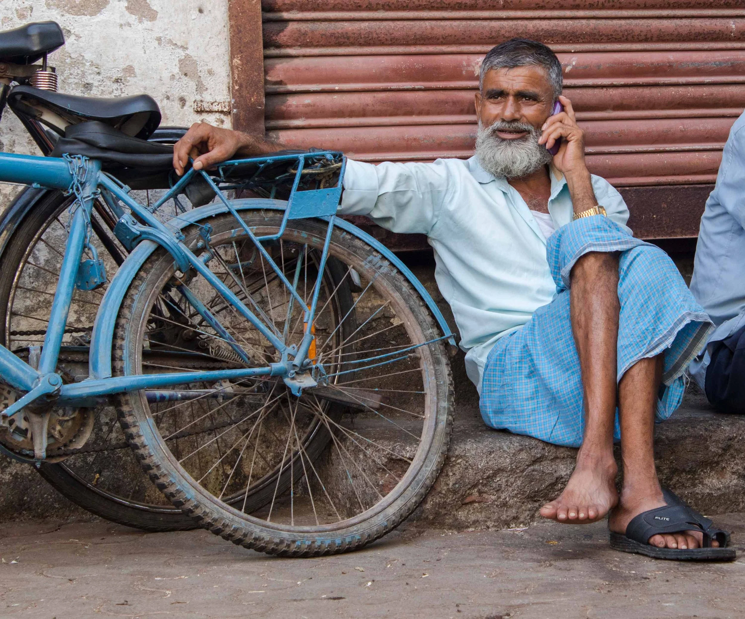 An older man in India with a gray beard, wearing a light blue shirt and blue pants, sitting on a street curb, talking on a cellphone, next to a blue bicycle. His legs are crossed with one shoe on and one off.