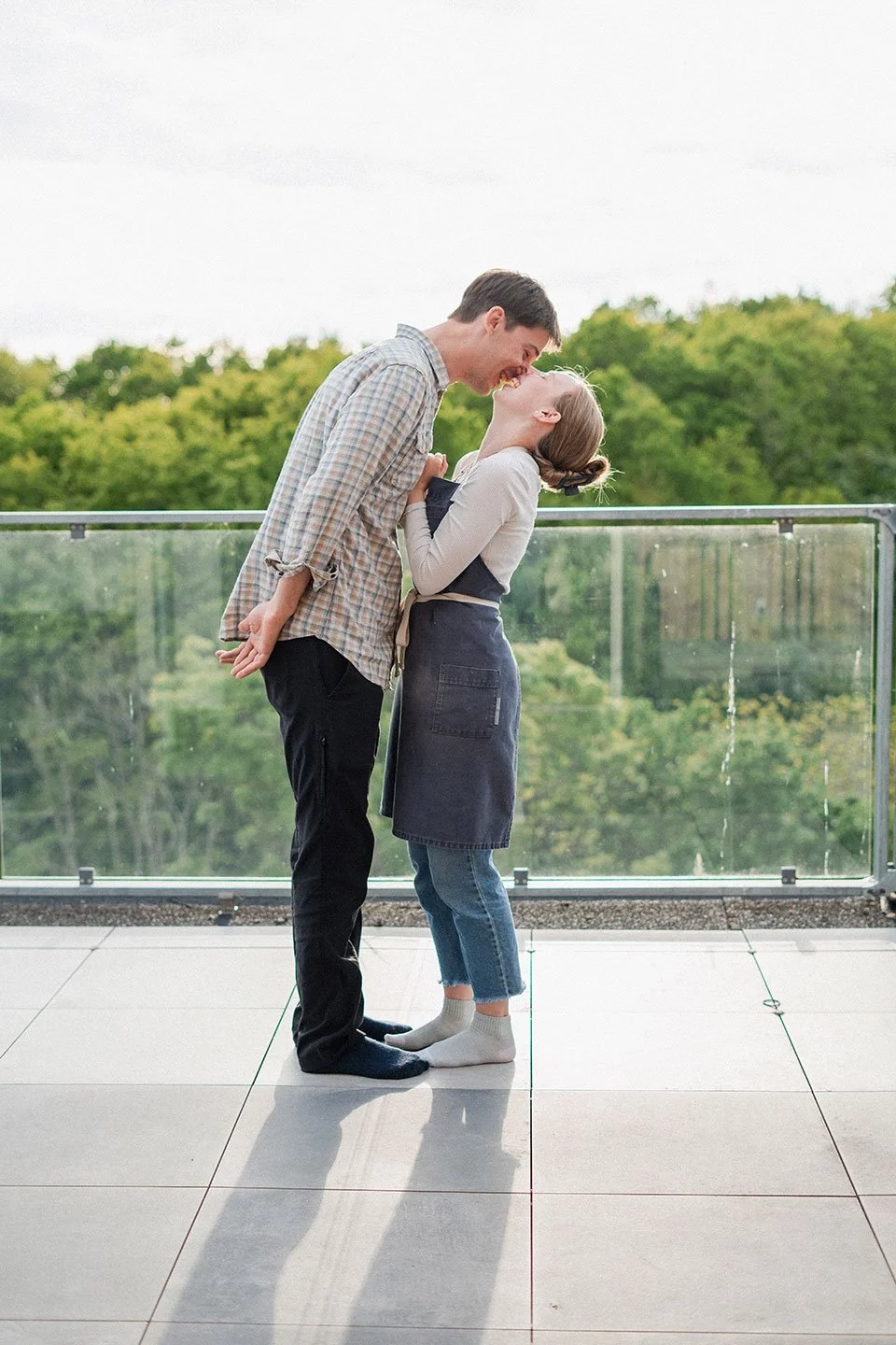 A couple is standing on a balcony, laughing and leaning into each other with their foreheads touching, surrounded by lush green trees.