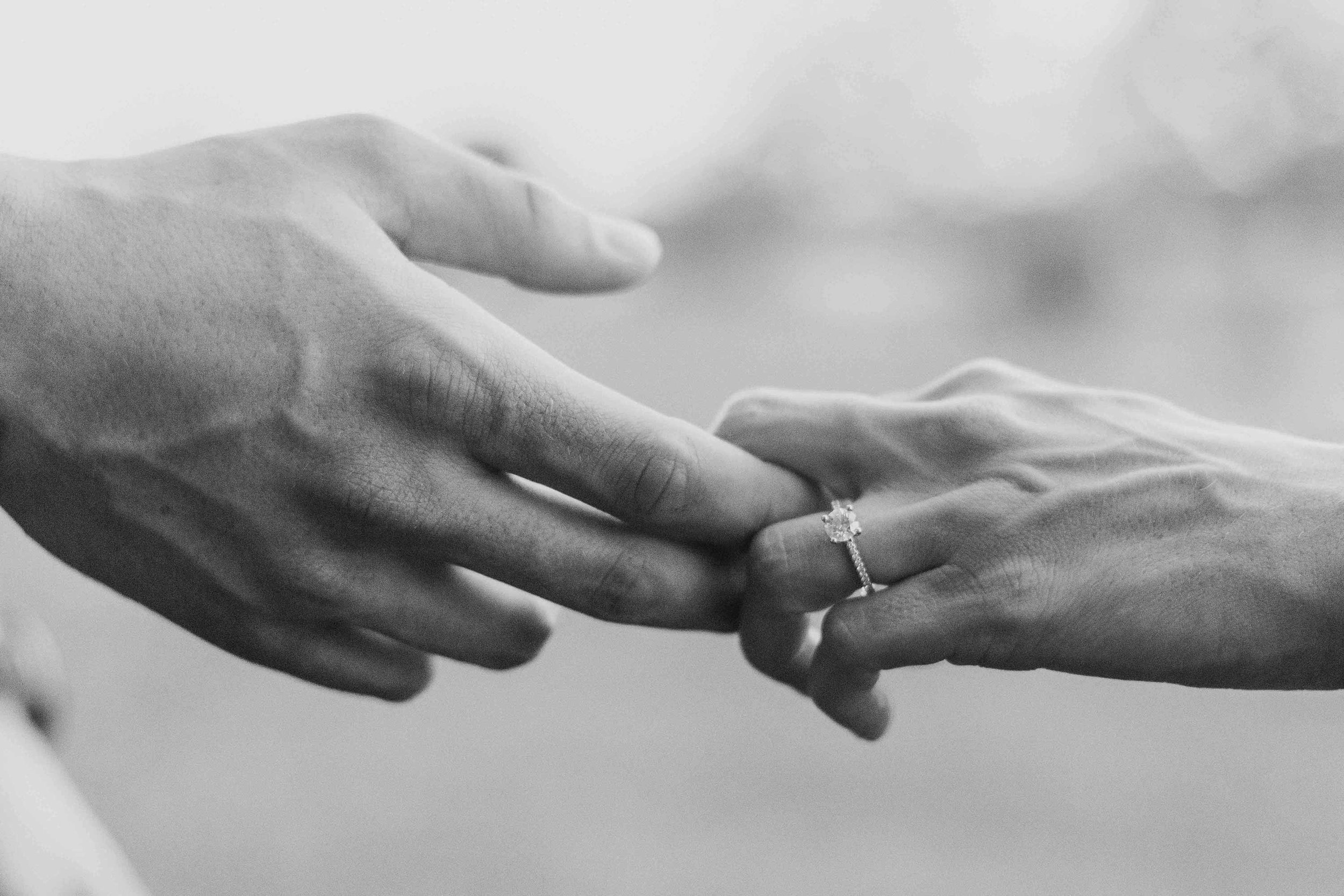 Close-up black and white photo of two hands with one wearing an engagement ring, gently touching