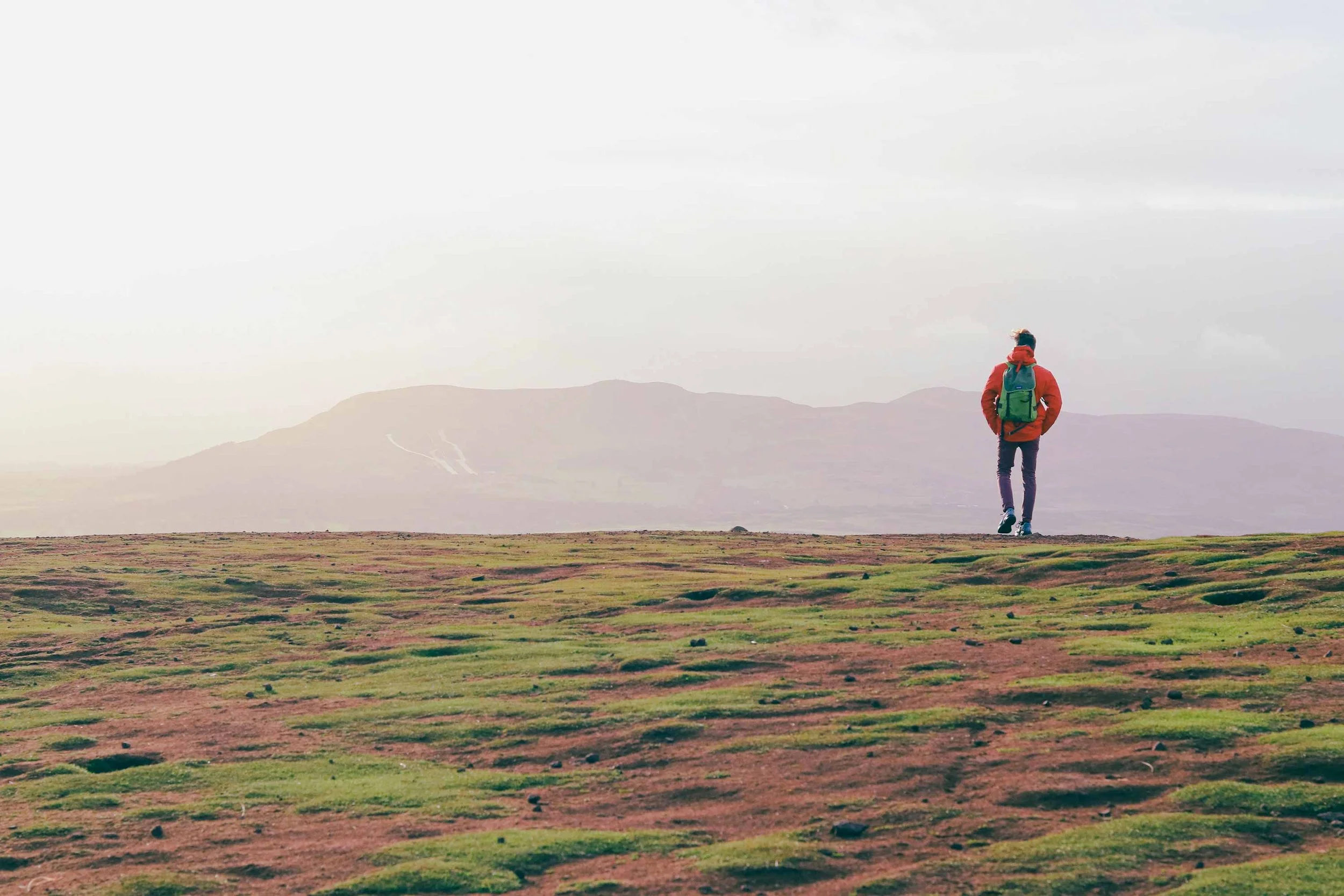 A person hiking in an open, grassy landscape with mountains in the distance on a cloudy day on Arthur's Seat in Edinburgh, Scotland.