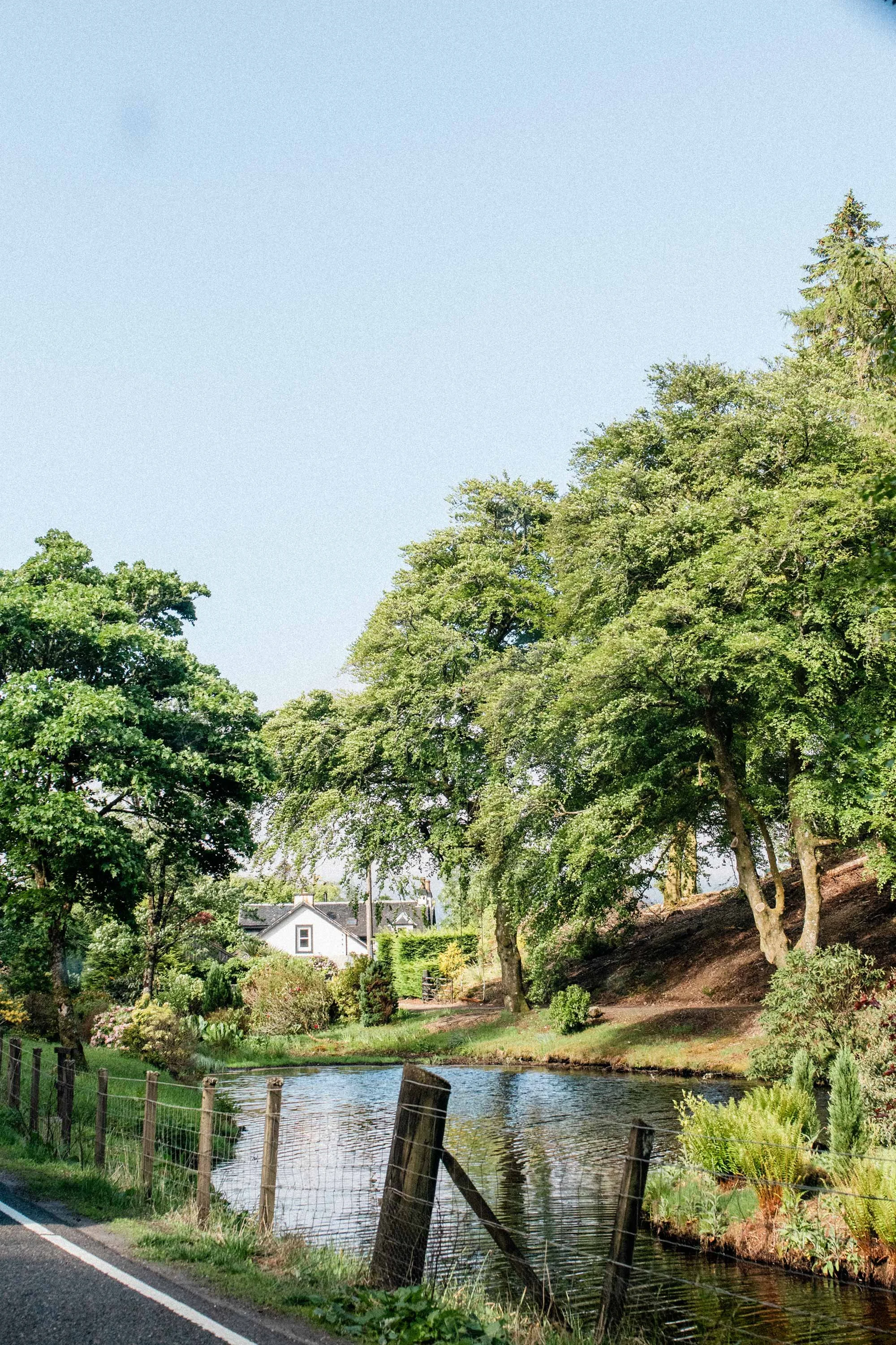 A peaceful, storybook-like photograph with a creek or small river running alongside a road, bordered by a wooden fence. There are lush green trees and a house under a clear blue sky.