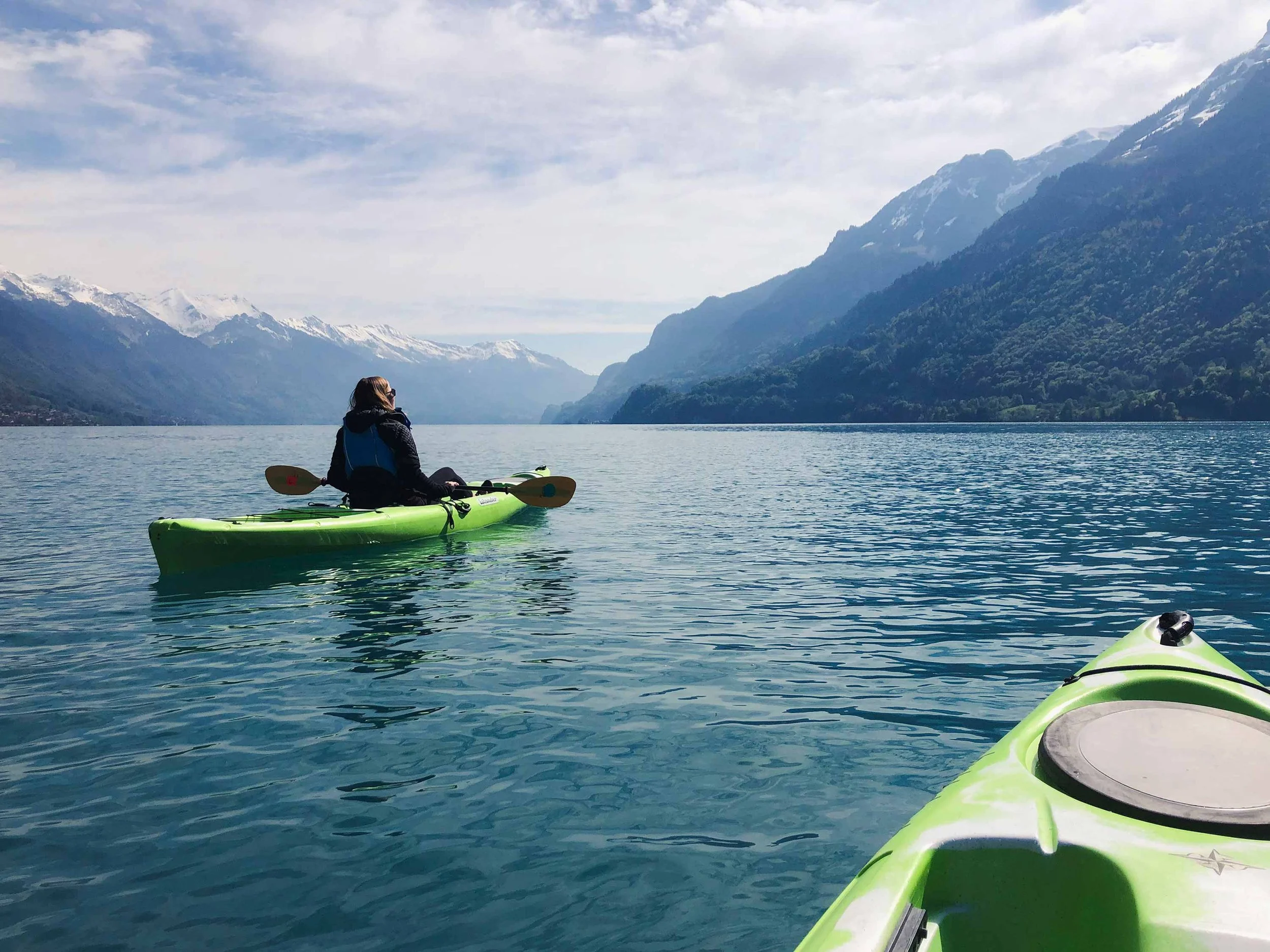 A person kayaking on a lake in the Swiss Alps surrounded by snowy mountains and forested hills under a partly cloudy sky.