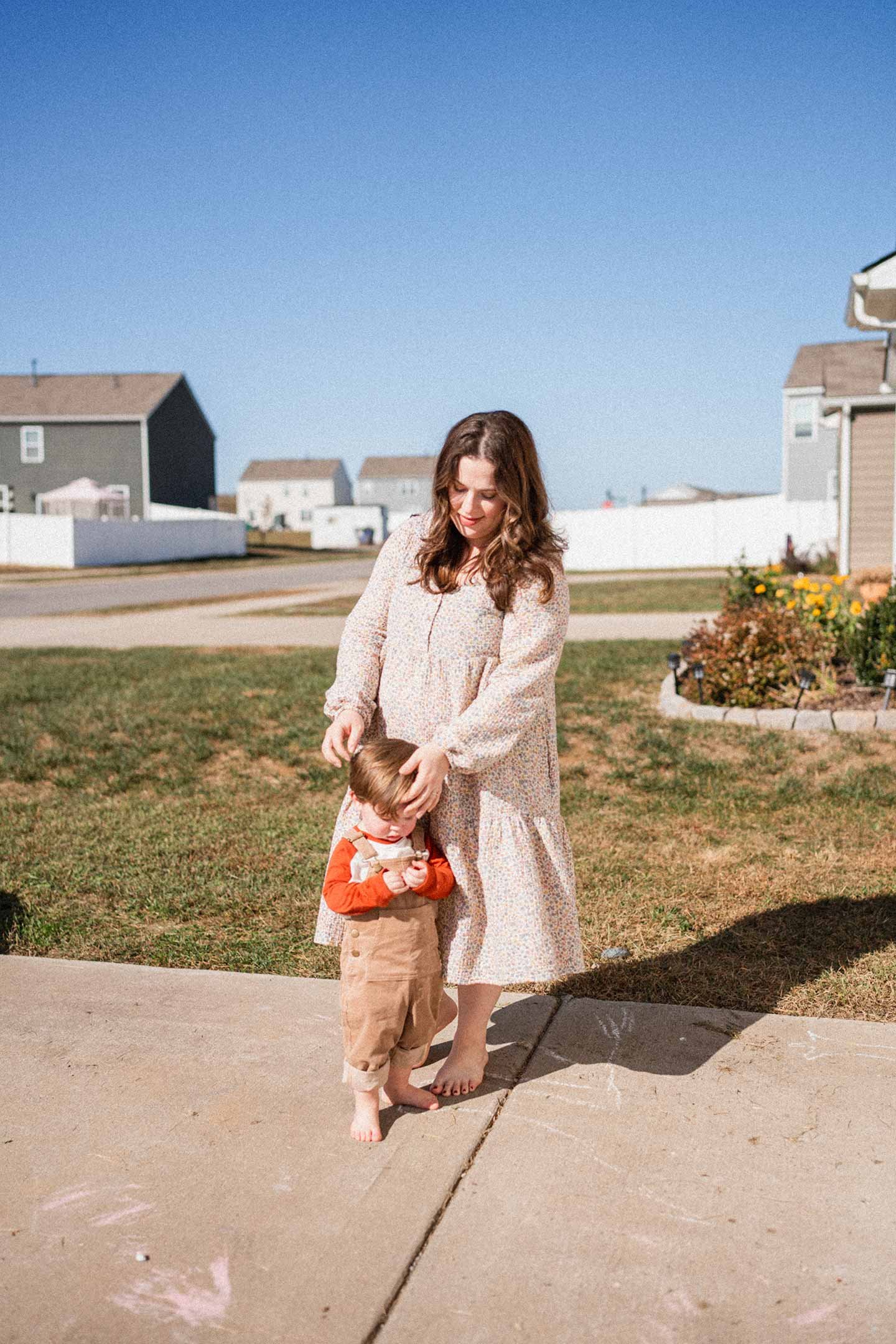 A mother stands with her little boy in their chalk covered driveway on a sunny day. She, in a long, flowing dress gently pats her son's, who wears an orange shirt and yellow overalls, head.