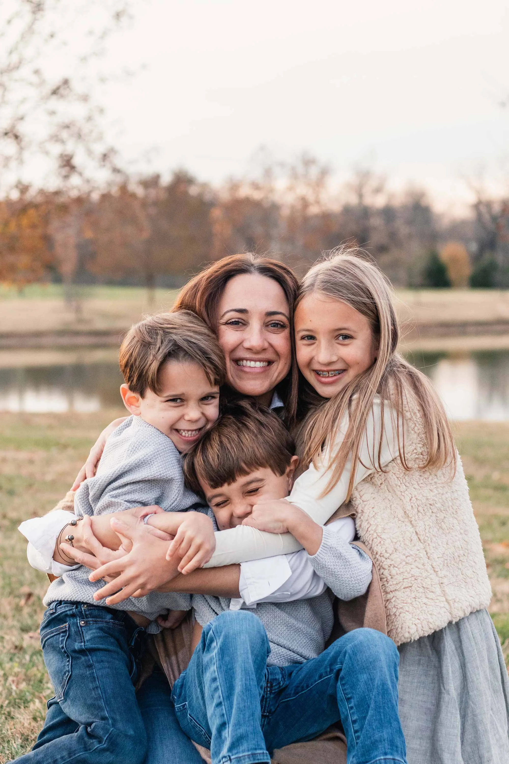 A mother and three children snuggle close in a bear hug outside near a lake.