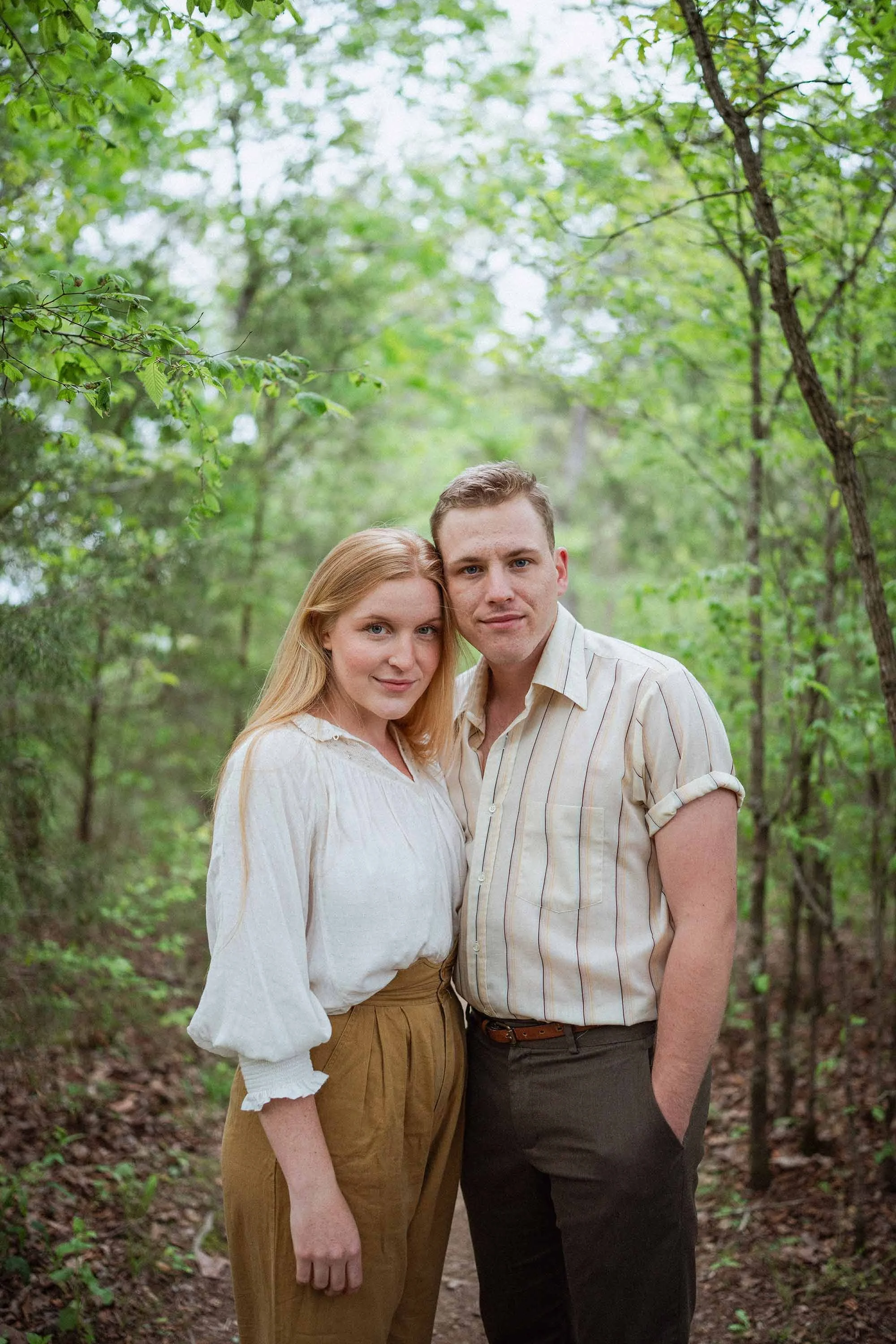 A young couple standing close together, framed by trees, looking at the camera. She has strawberry blond hair, a white puffy shirt, and yellow pants. He has a striped shirt and green pants.