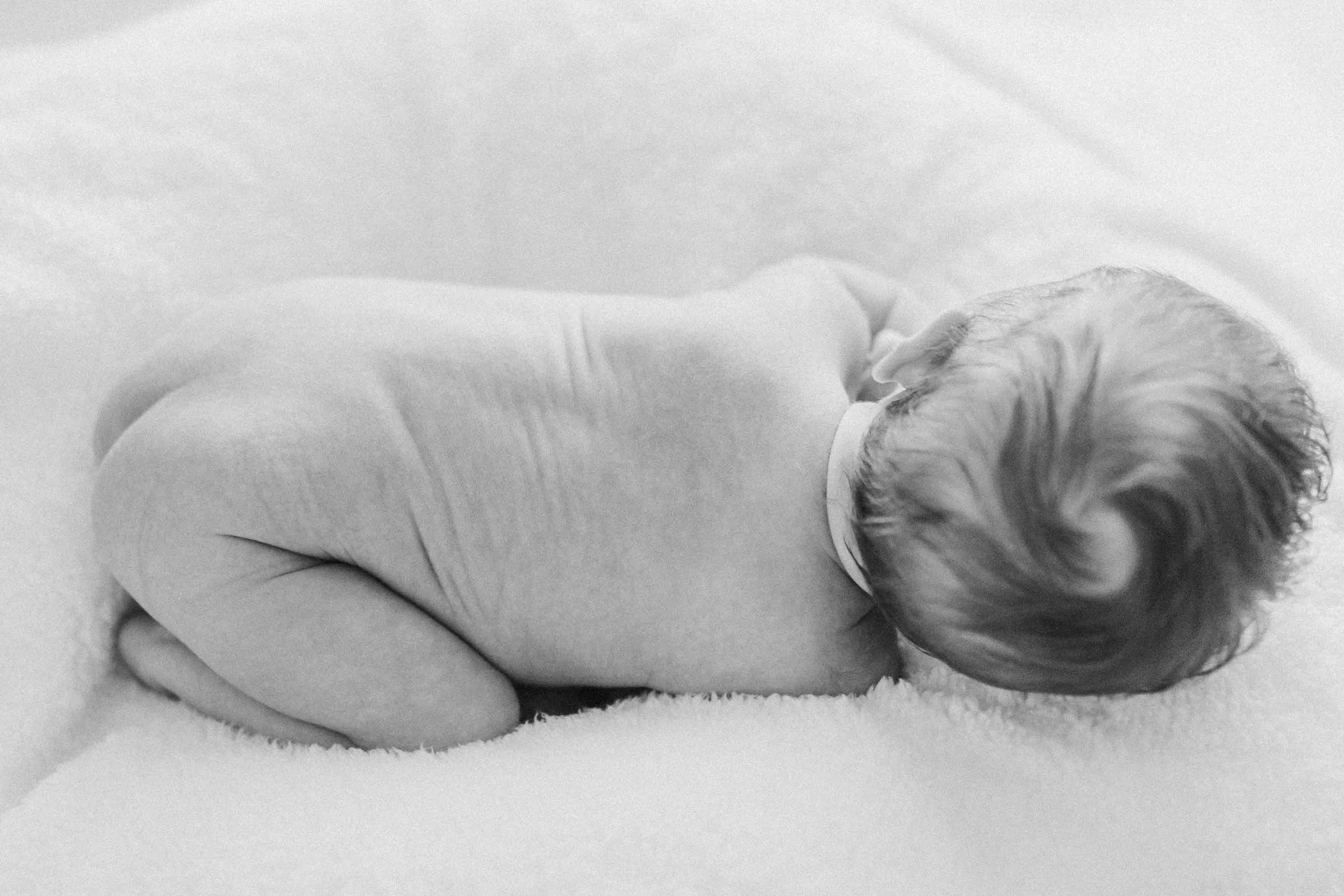 In black and white, from above, a naked sleeping newborn baby lying on a soft blanket, showing her hair, back, and tiny body.