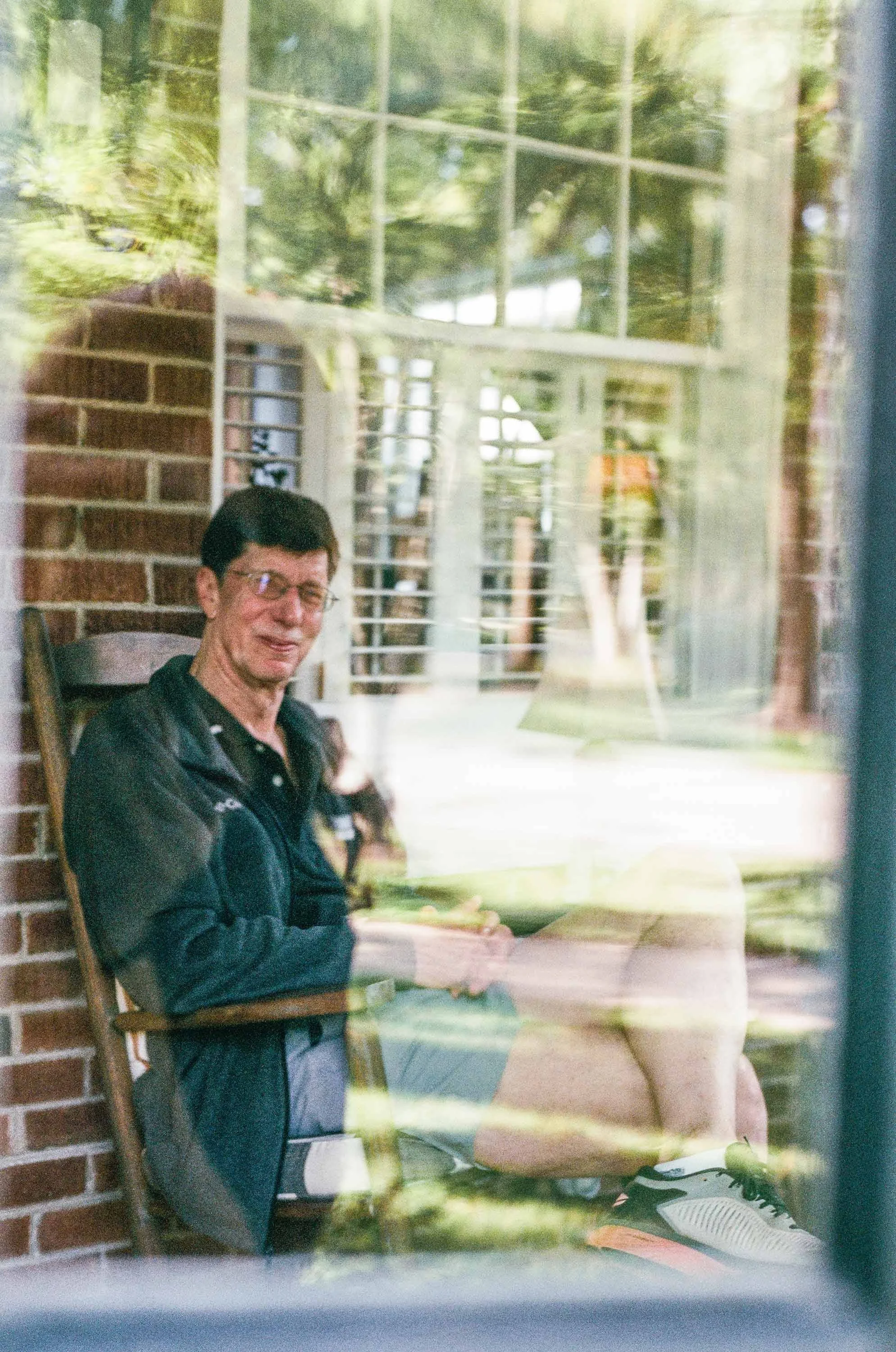 Color film photo shot through a window of a man sitting in a sun room. The man sits smiling with his legs crossed. You can see the reflection of the trees against the window.
