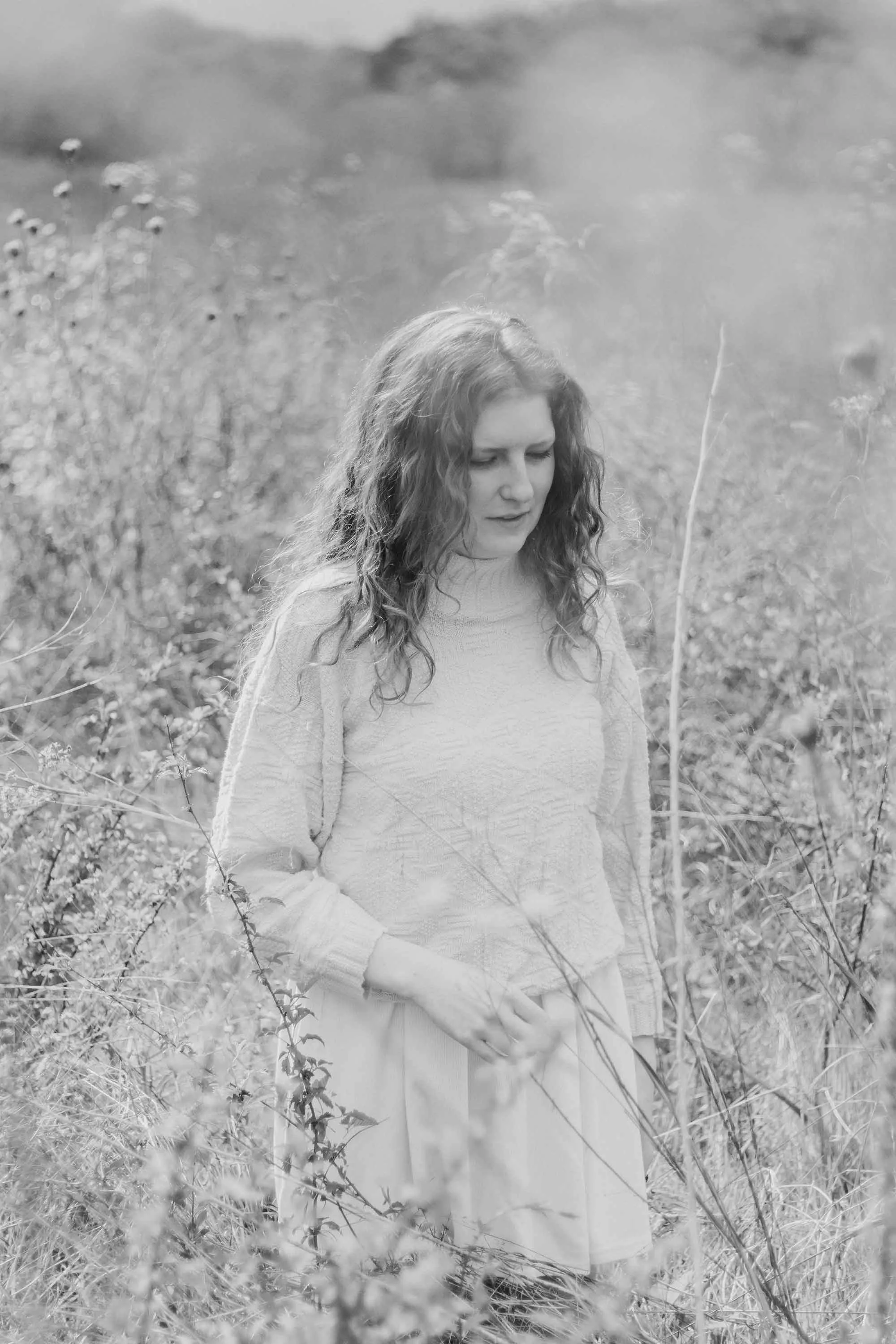 A young woman wearing a sweater and a skirt, standing in a field with tall grass and wildflowers with a blurred foreground.