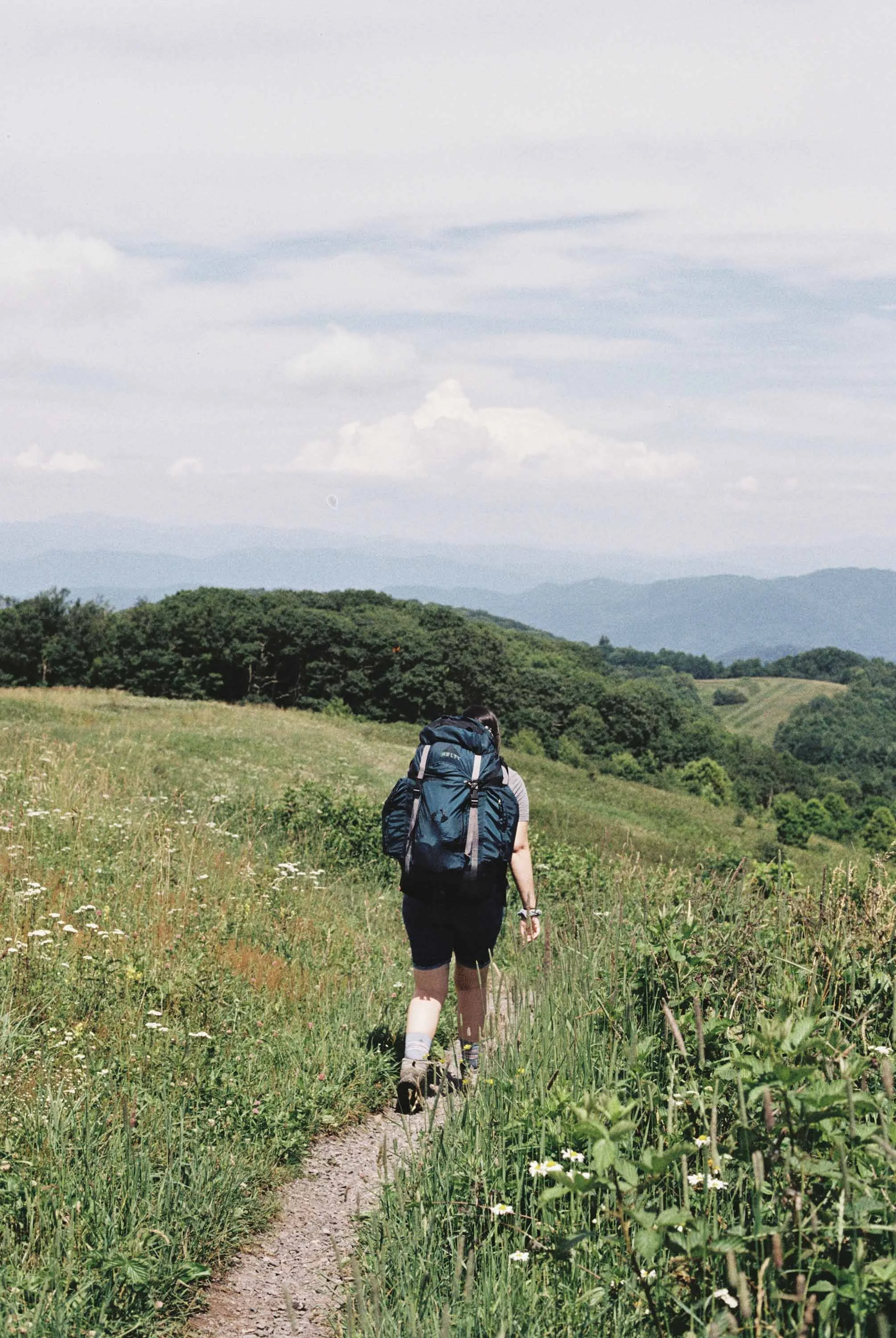 A woman hiking in the Smokey Mountains with a backpacking pack on a narrow trail through a grassy field with wildflowers, green hills, and mountains in the background under a partly cloudy sky.