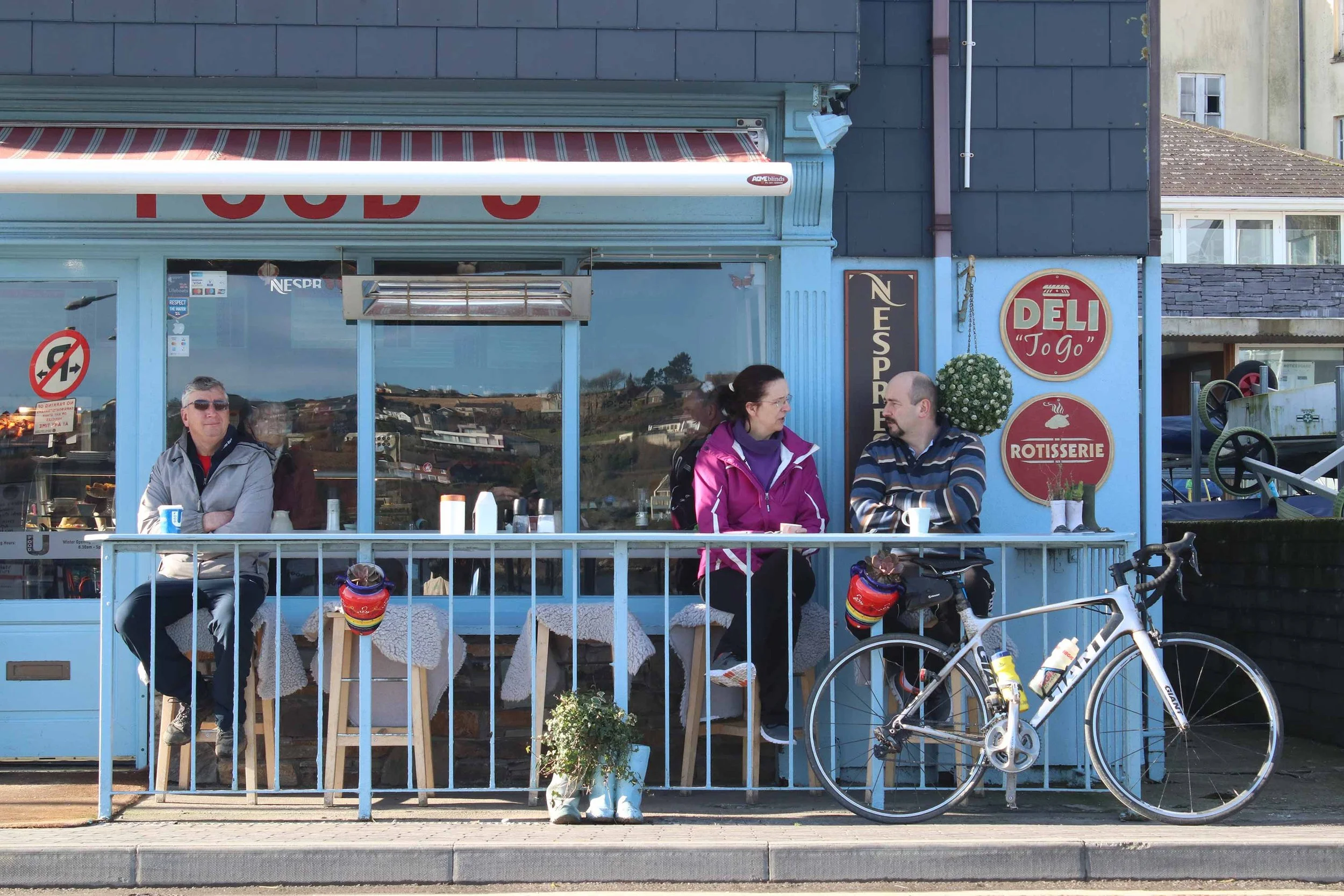 Three people are sitting outside a blue café with a bicycle parked nearby in Kinsale, Ireland. A man and woman talk together, a man sits alone, and a woman looks out the window.