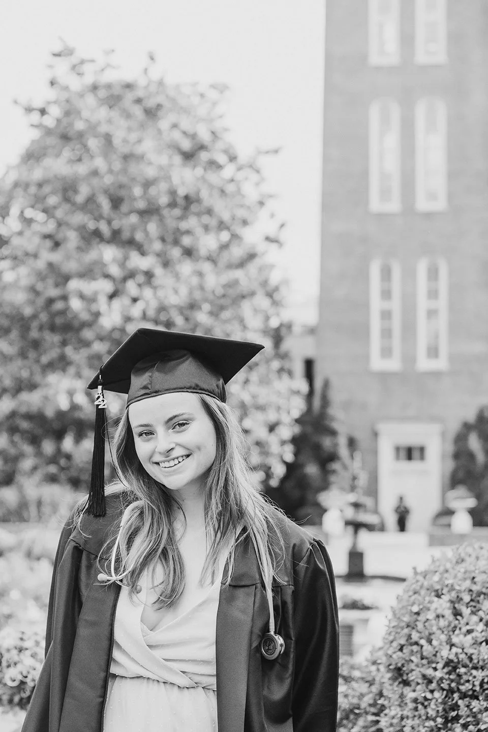 A black and white photo of a young, woman, nursing graduate in graduation cap and gown smiling outdoors with the Belmont bell tower blurred in the background.
