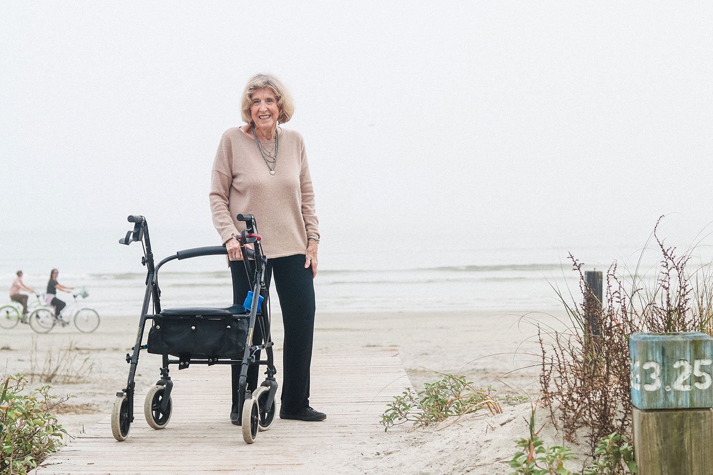 An elderly woman standing on a beach path with a walker, smiling at the camera, in front of the ocean.