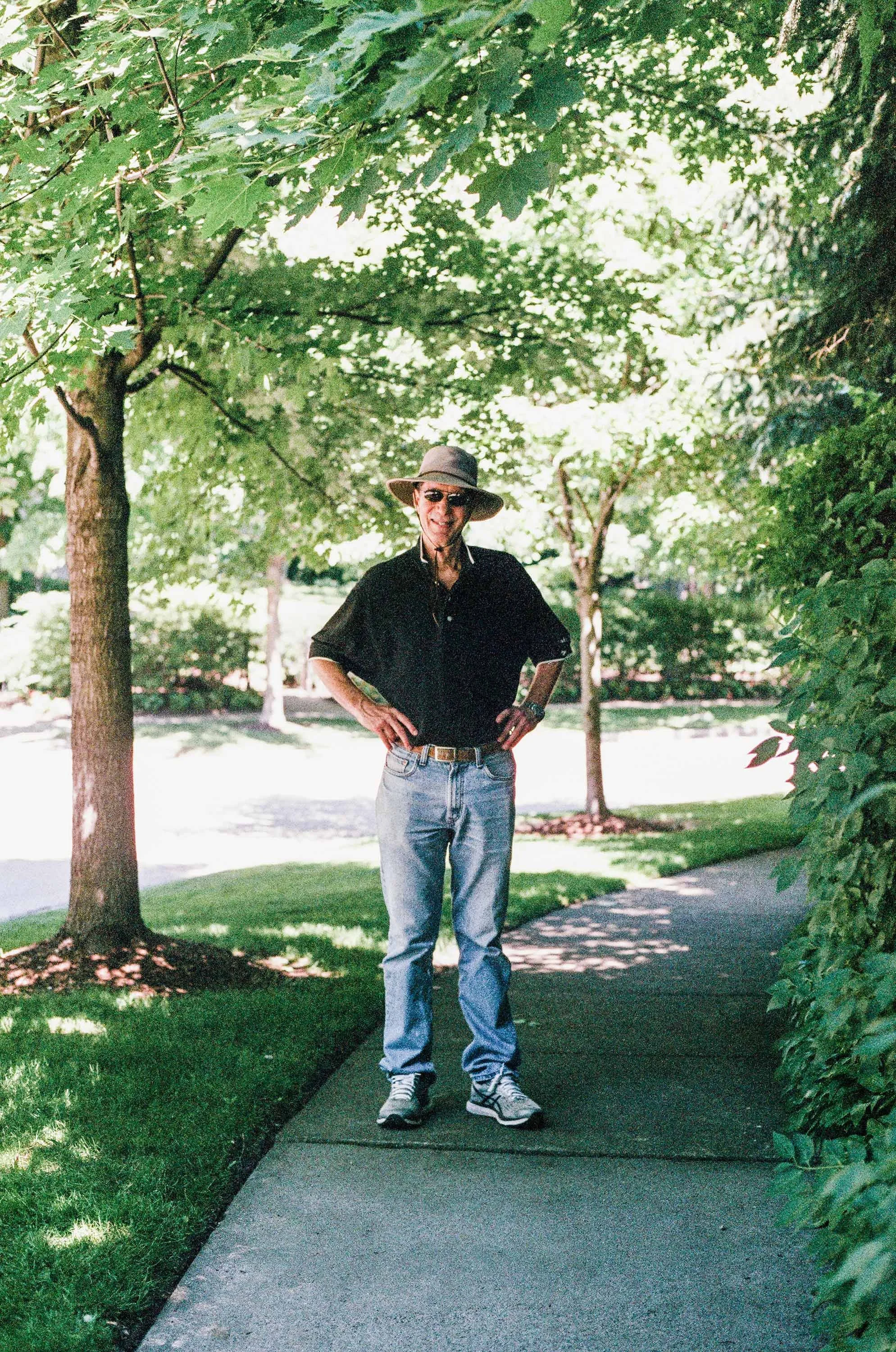 Color film photo of an older man standing on a sidewalk, wearing sunglasses, a wide-brimmed sun-hat, a black shirt, jeans, and sneakers, with trees and greenery around him.