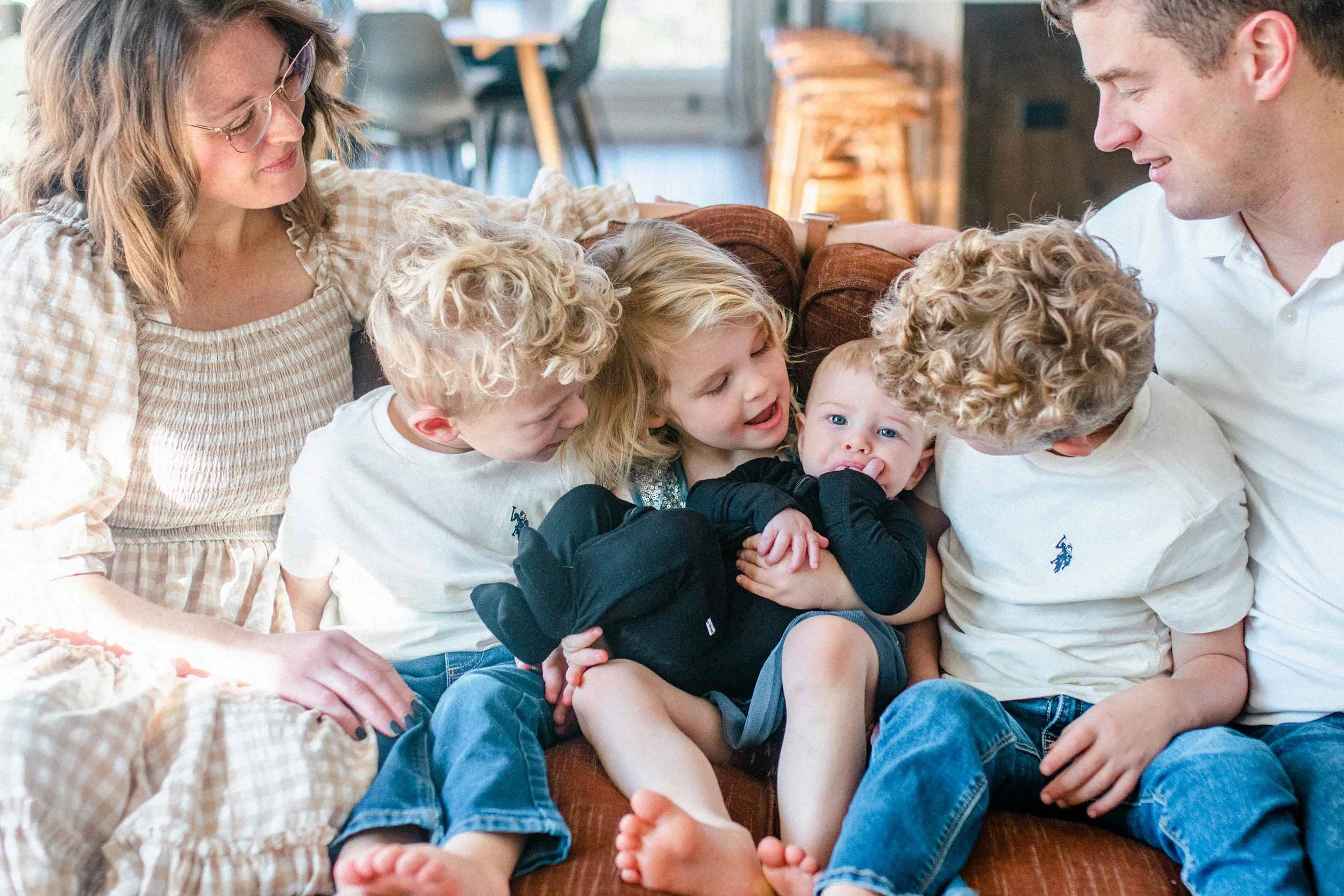 Four young kids which curly blond hair sit on a couch together, sandwiched between their mom and dad. All of them look at the baby while the baby looks at the camera.
