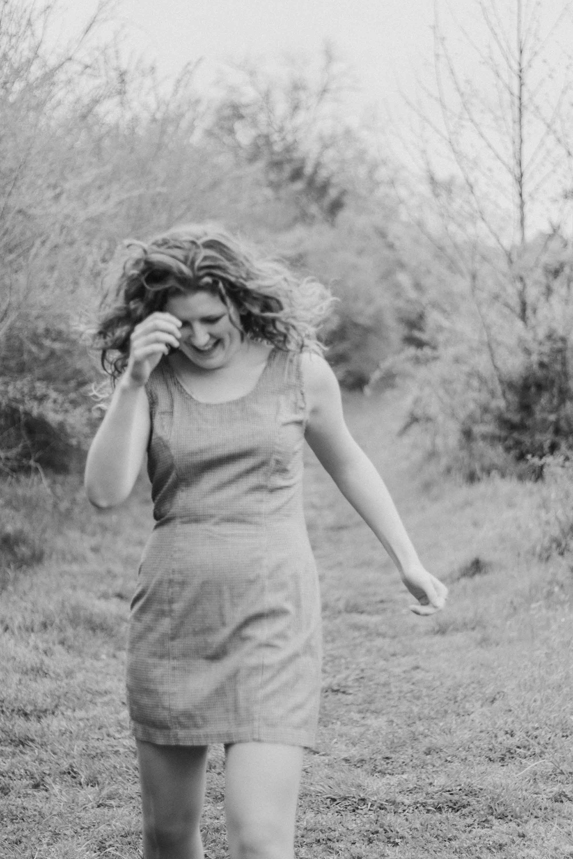 A slightly blurred, black and white photo of a woman smiling and walking outdoors on a grassy path surrounded by trees.