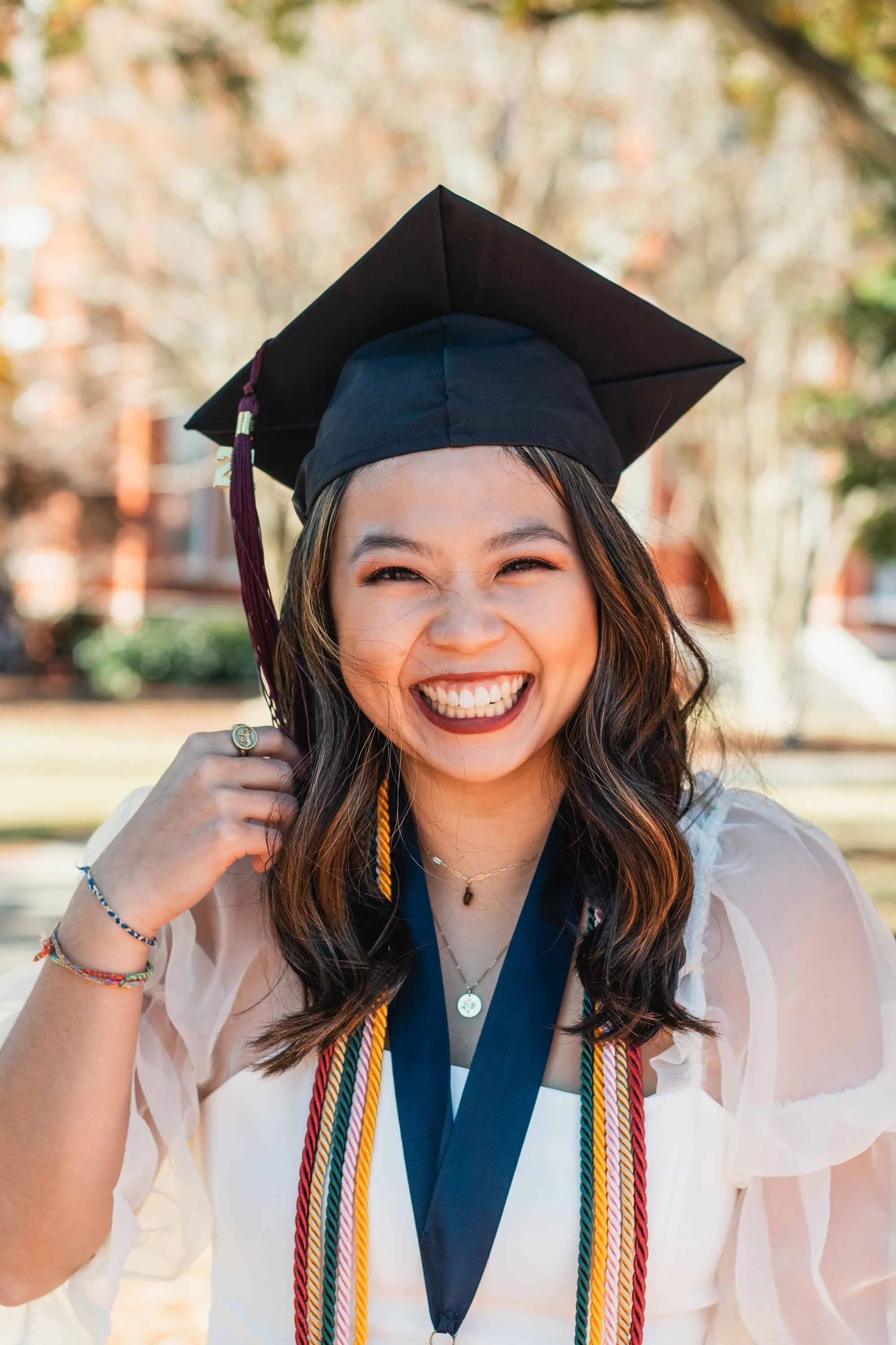 A young woman with a big smile in a graduation cap and gown outdoors on a sunny day.