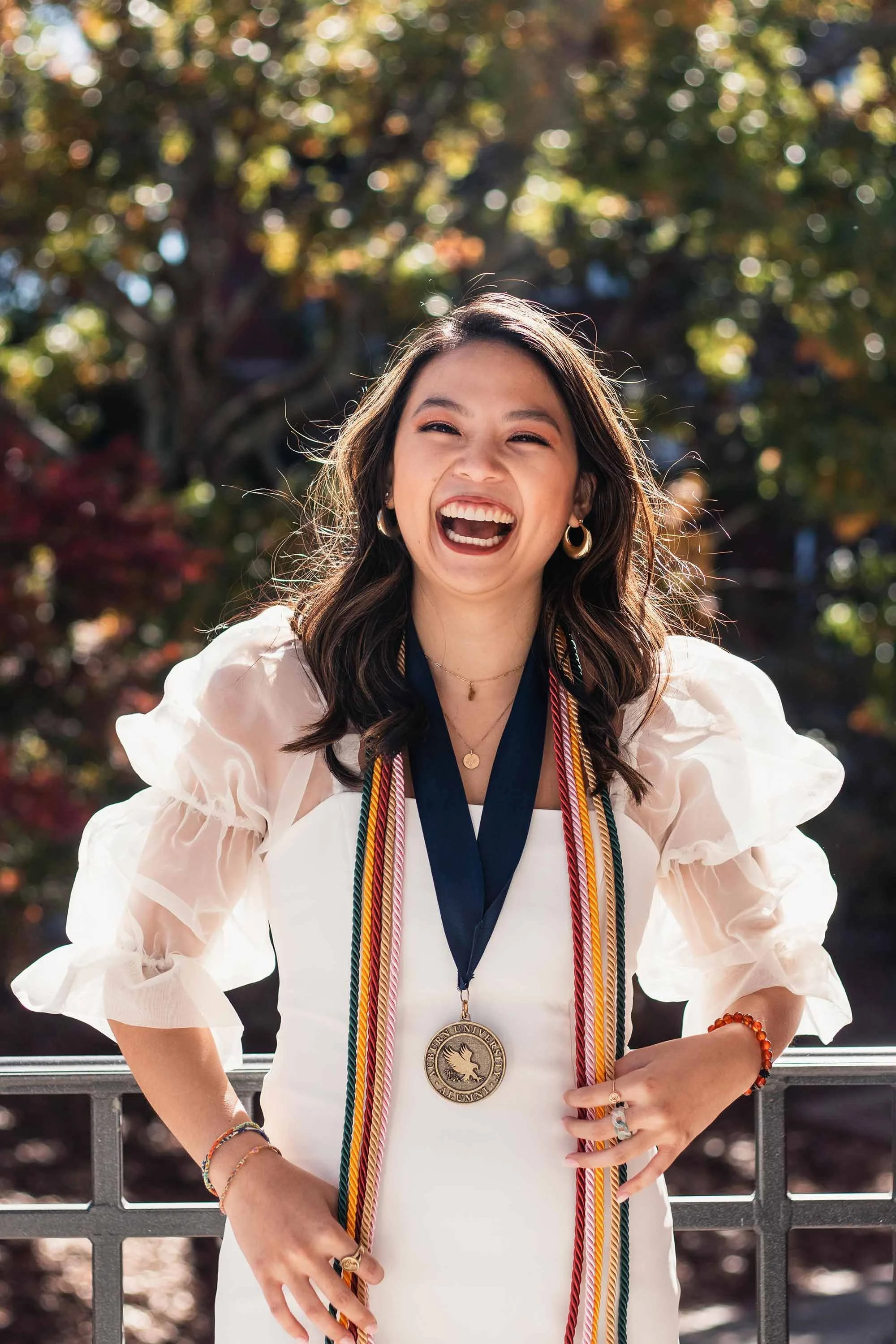 A young woman celebrating graduation outdoors, wearing a white dress, with graduation cords and a medal around her neck, smiling and laughing.