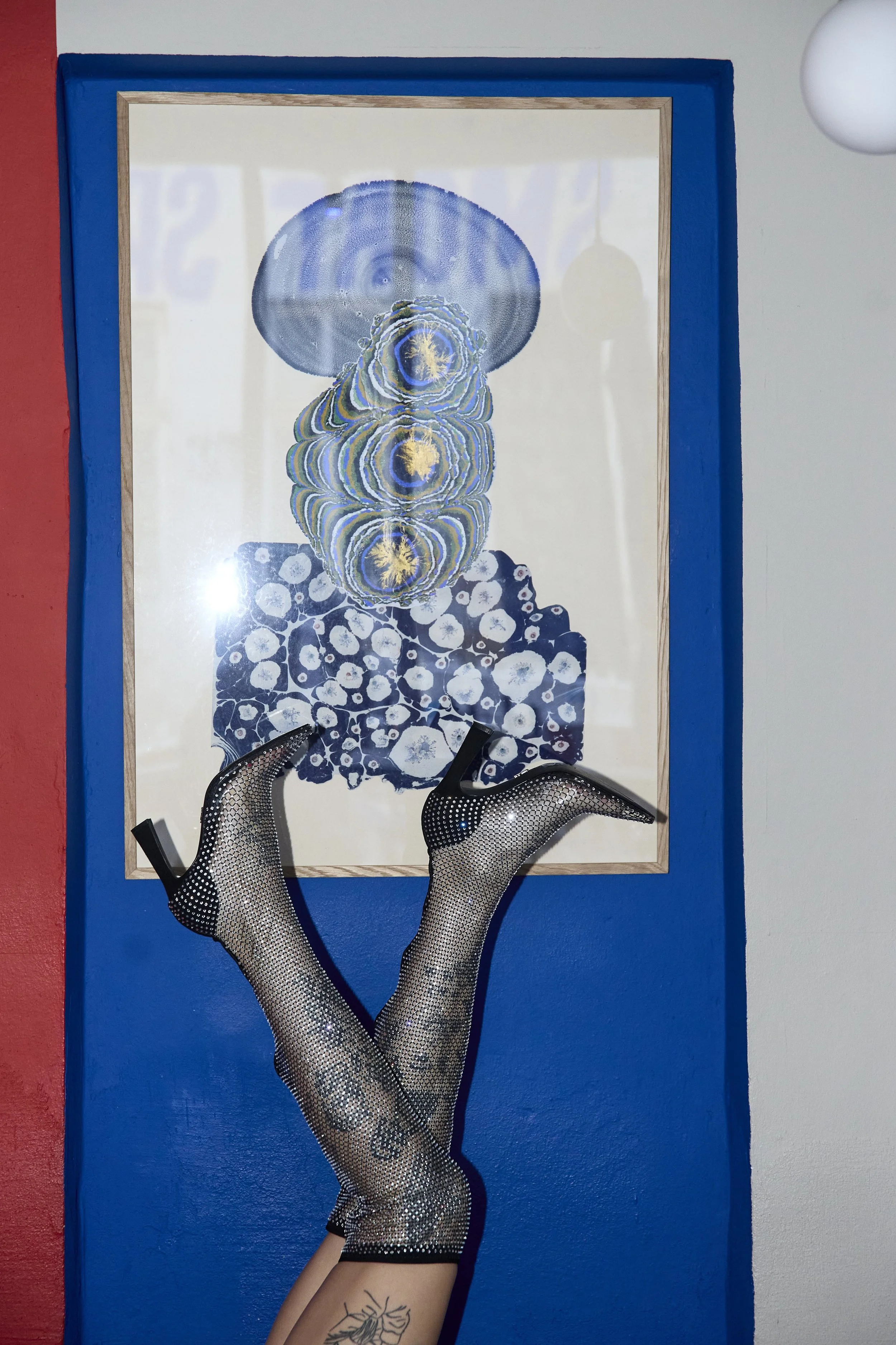 Framed collage abstract artwork by Gökce Tercioglu displays concentric circles in shades of blue, yellow, green and gold with a person's legs in fishnet sparkly stockings and high-heeled shoes in front of it, and part of their tattooed legs visible.