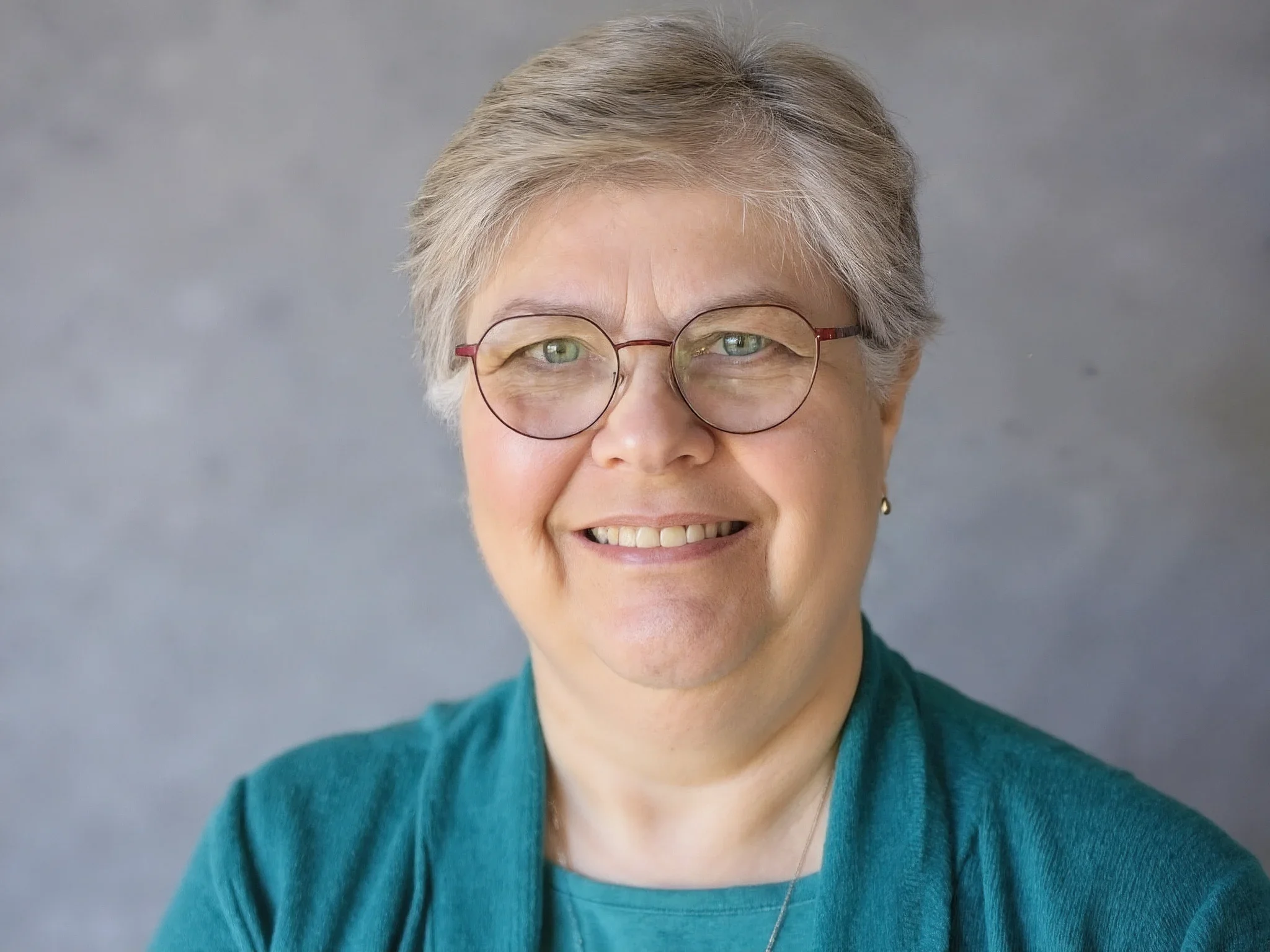A smiling woman with short gray hair wearing glasses, a teal shirt, and a matching teal cardigan against a gray background.