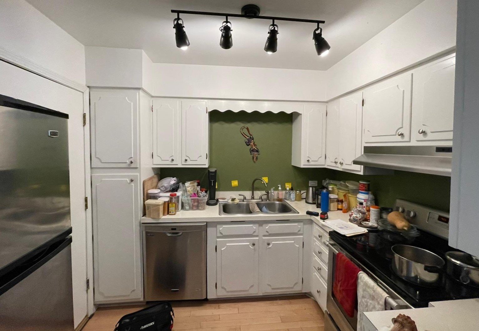 A cluttered kitchen with white cabinets, a green wall behind the sink, and various kitchen items on counters and stove.