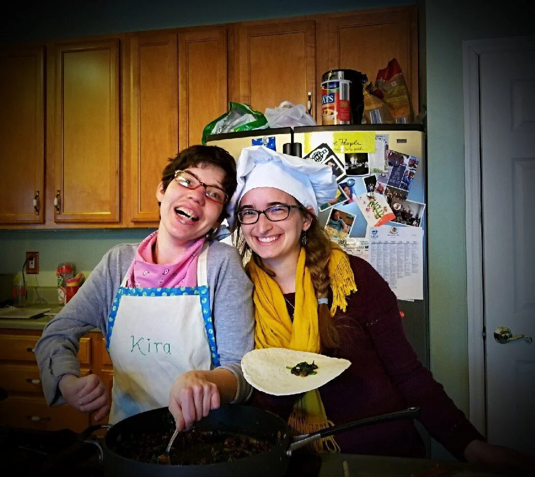 Two women smile in a kitchen, leaning into each other. One wears an apron and is stirring a pot, the other is wearing a chef's hat and holding food.