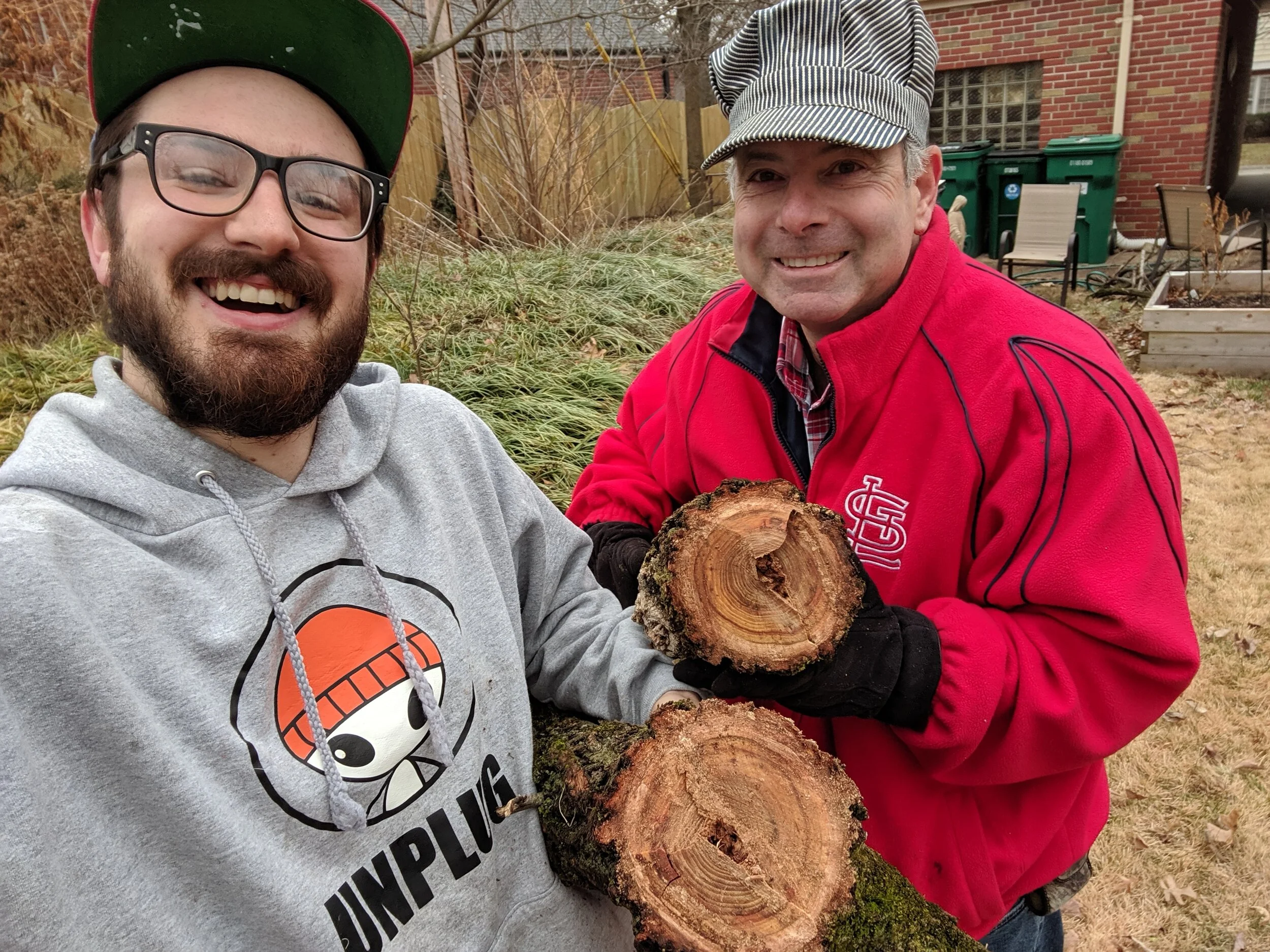 Two men smile at the camera while holding logs of wood. They are outside, standing in grass, with a brown fence in view. One man is wearing a red sweatshirt; the other is wearing a gray hoodie.