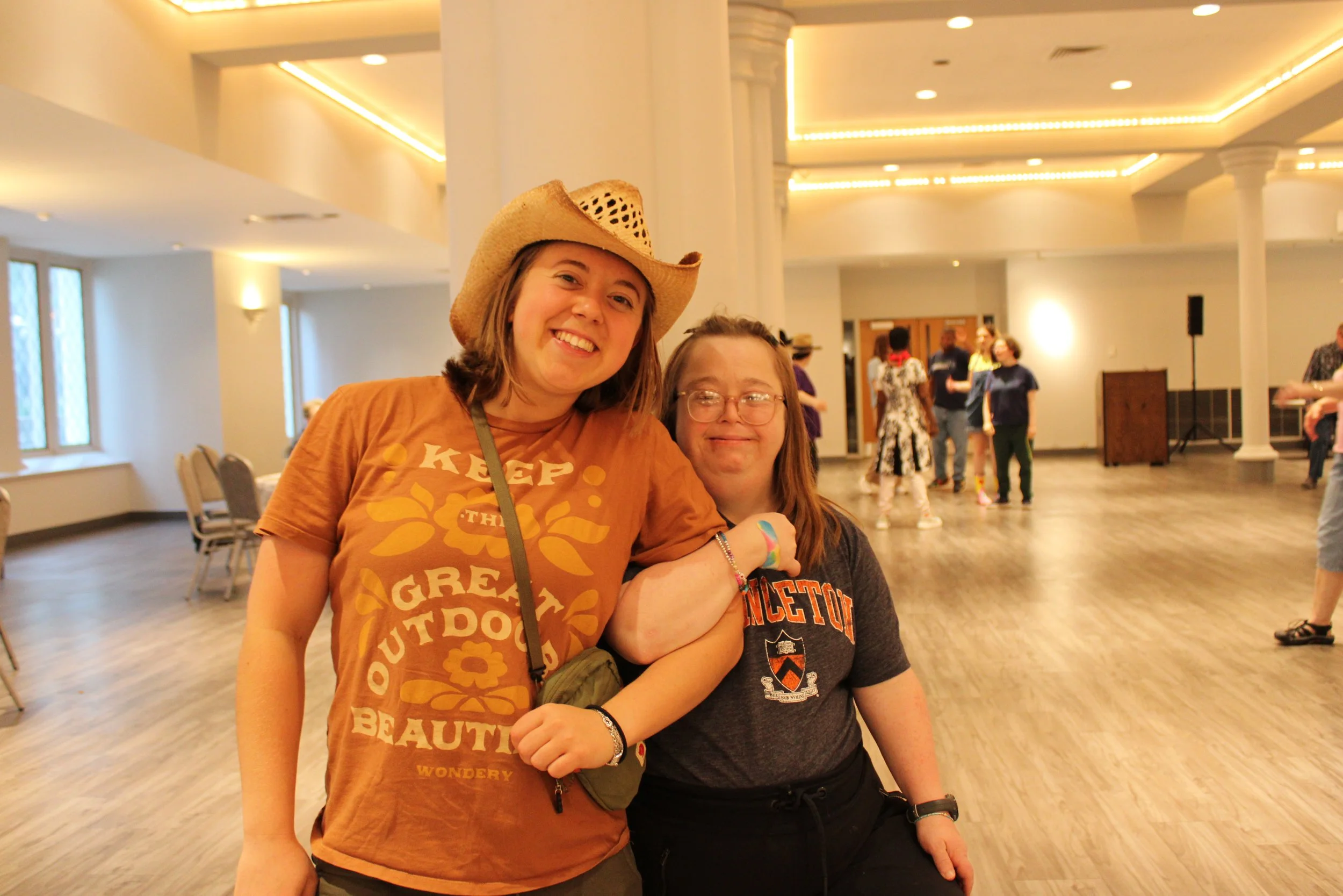 Two women link arms and smile into the camera, one wearing a cowboy hat.
