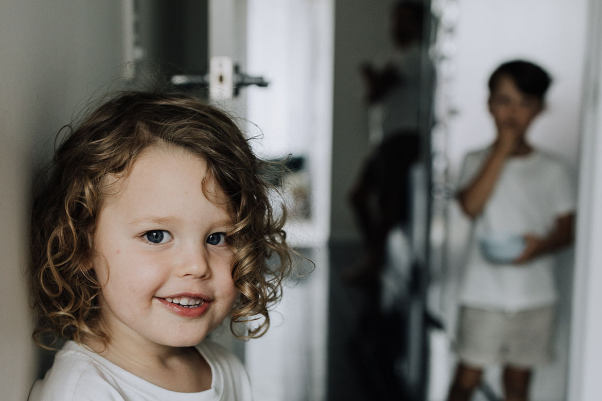 A young child with curly hair smiles in the foreground while an older child stands softly out of focus in the background of a family home.