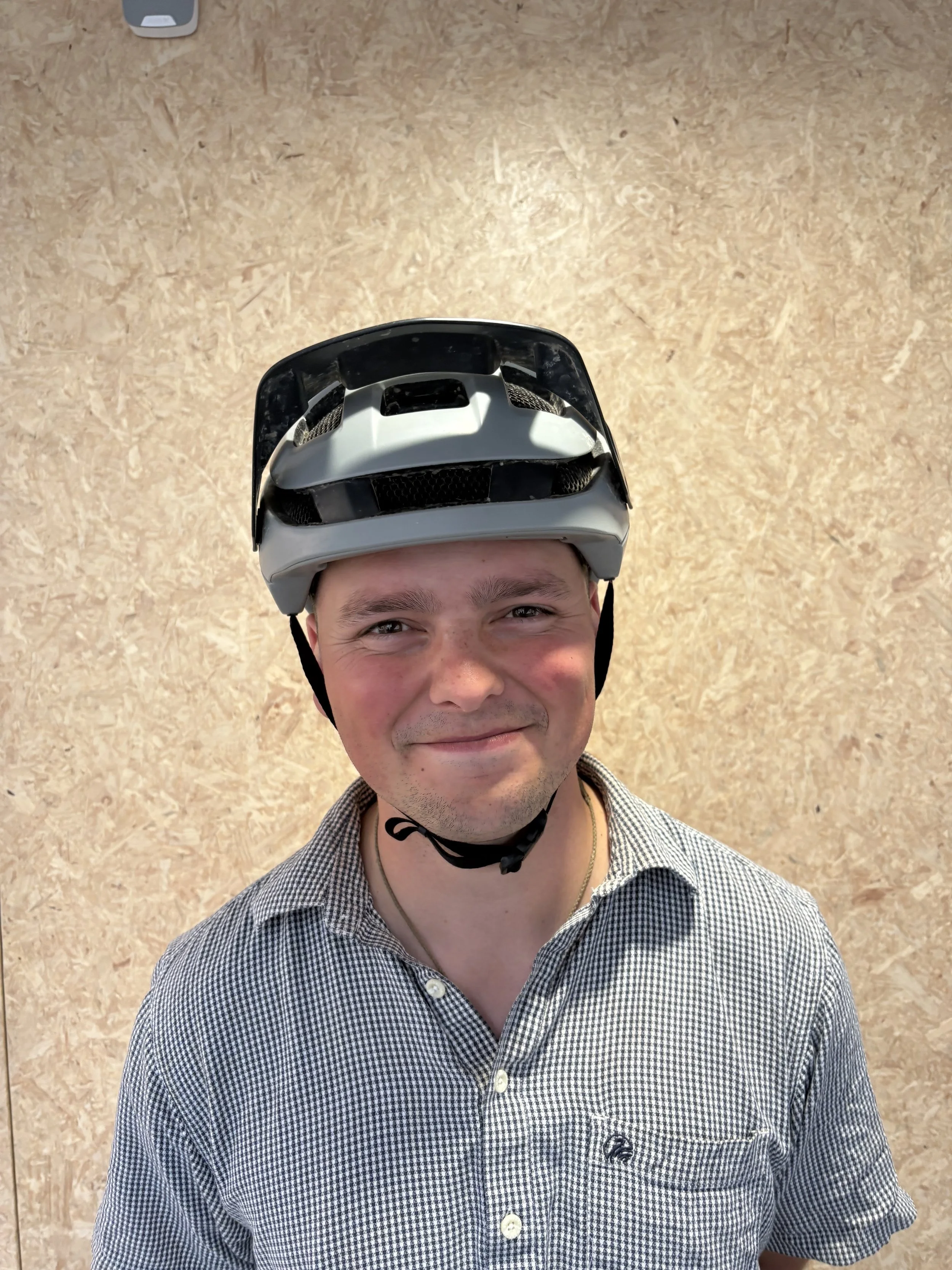 A smiling man wearing a bike helmet and a checked button-up shirt, standing against a light wood textured background.