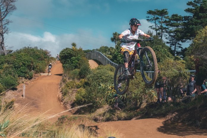 Child riding a mountain bike in mid-air during a jump on a dirt trail, with other riders and spectators in the background, surrounded by trees and bushes on a sunny day.