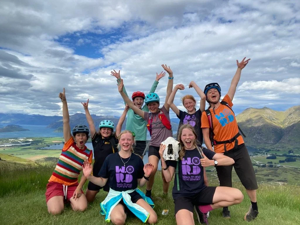 Group of young people wearing helmets and outdoor gear, smiling and celebrating on a hillside with mountains and a lake in the background.