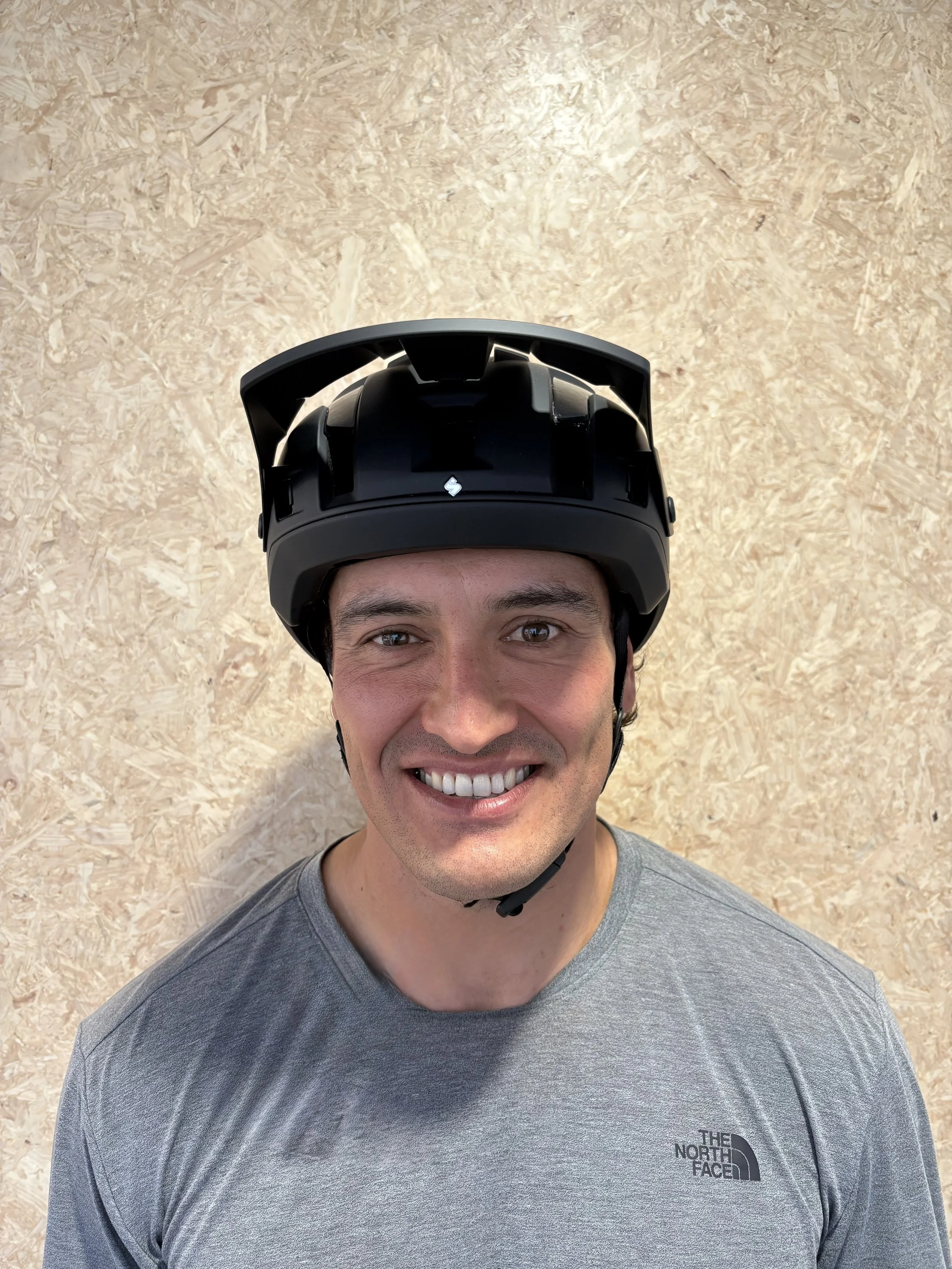 A smiling man wearing a gray T-shirt and a black helmet, standing in front of a beige wooden wall.