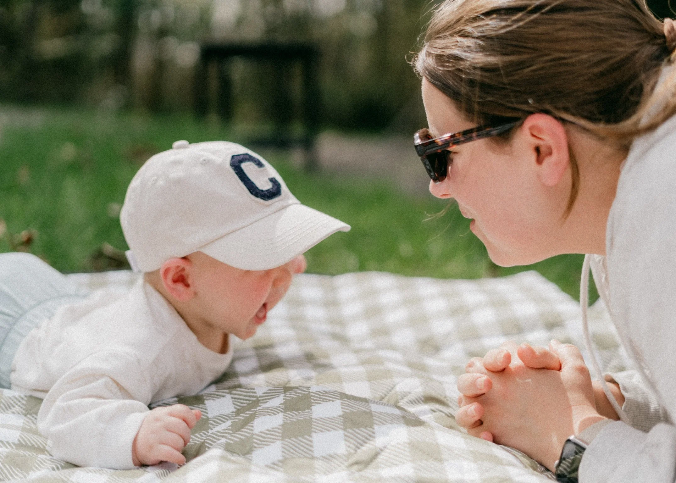 A woman and a young child are lying face to face outdoors on a blanket. The woman is wearing glasses and has brown hair, while the child is wearing a white baseball cap with a blue letter 'C' on it and a white shirt. They are closely gazing at each other with their hands clasped together.