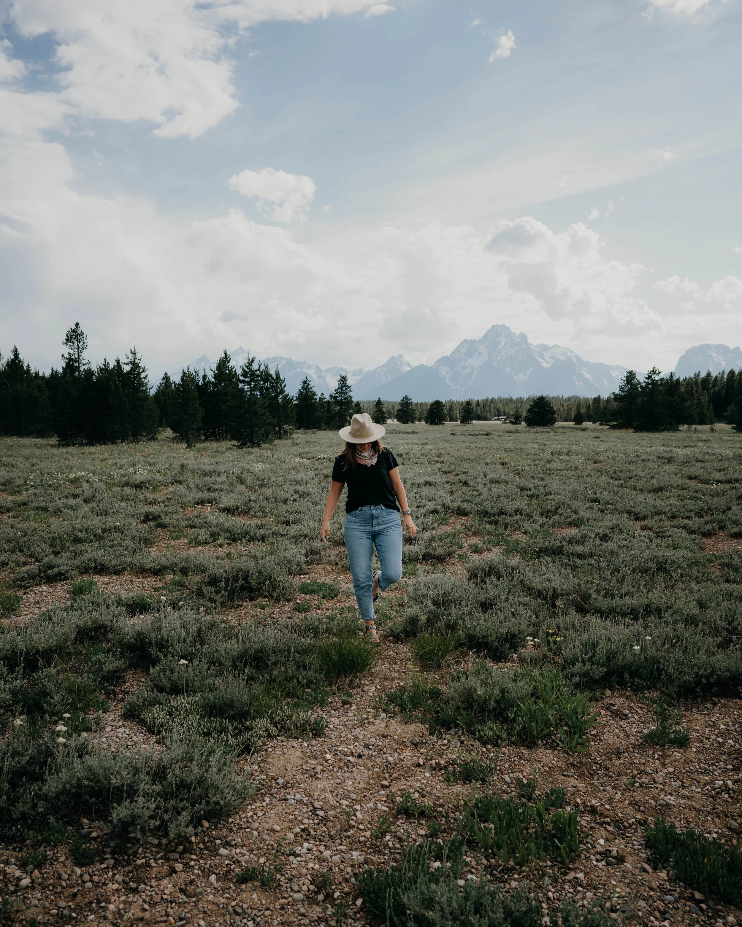 A woman in a hat and black t-shirt walking through a field of low bushes near a mountain range, with partly cloudy sky overhead.