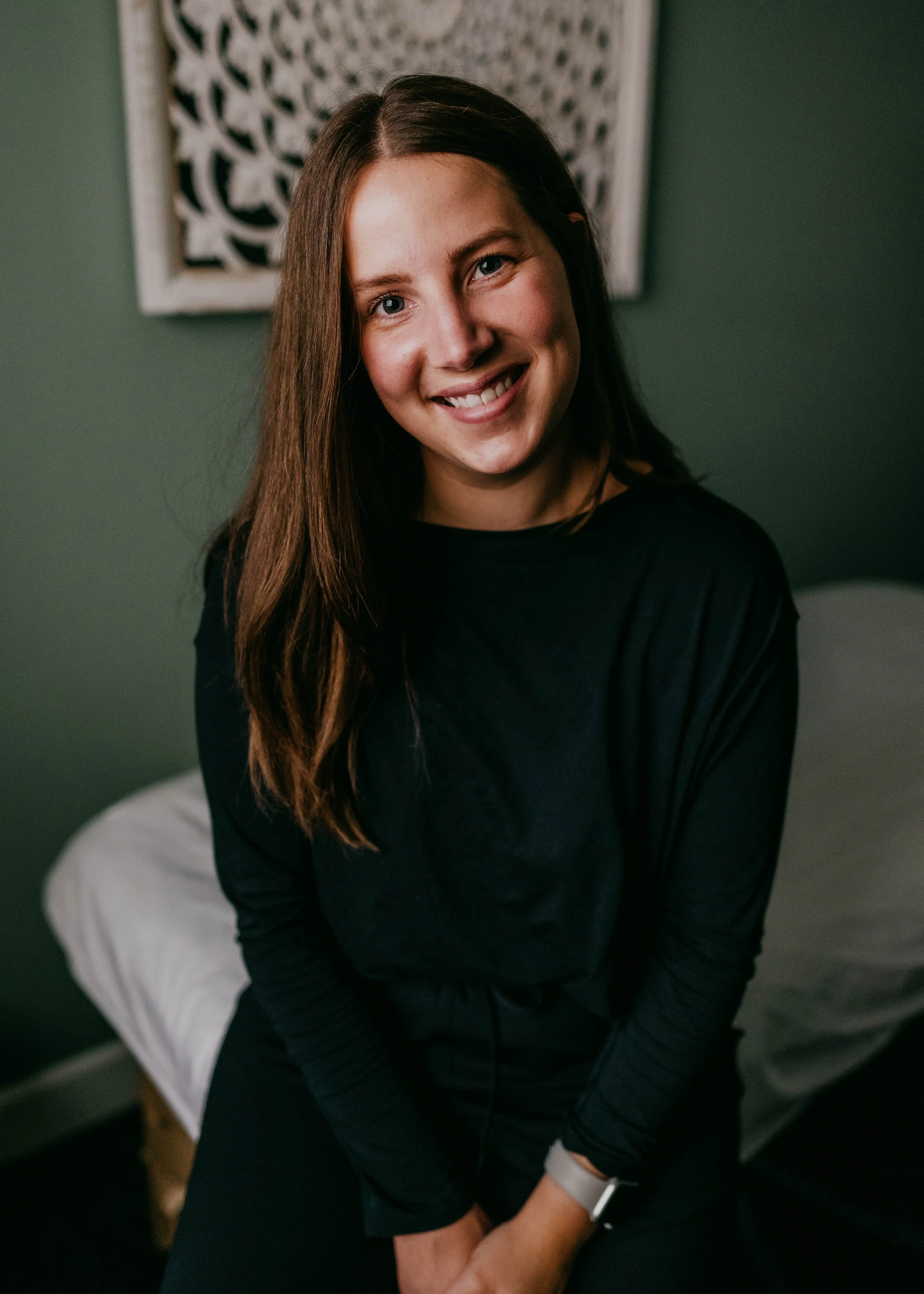 A young woman with long brown hair and blue eyes smiling, sitting on a bed with white sheets, wearing a black top and black pants, in a room with dark green walls and a decorative wall hanging.