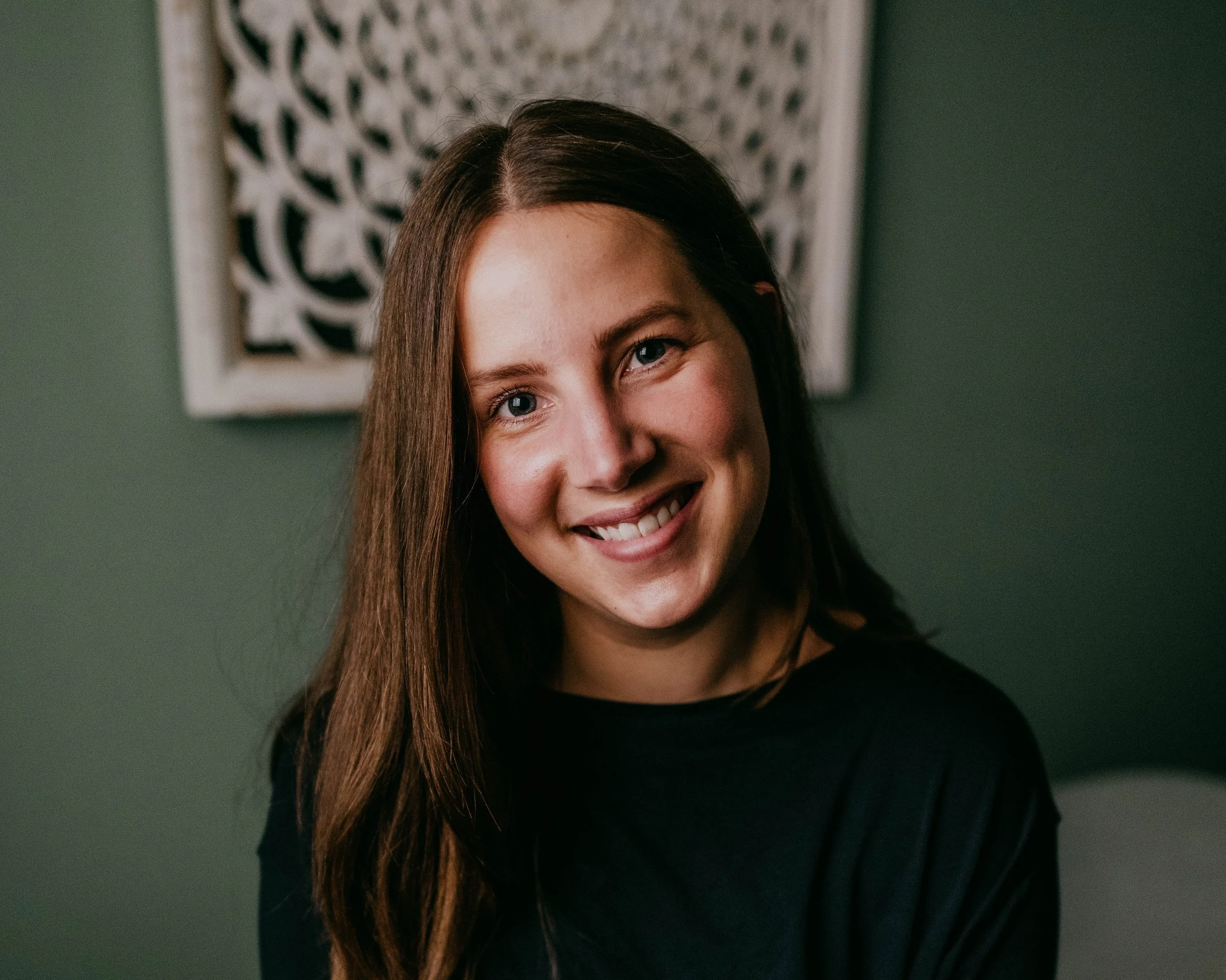 Close-up portrait of a young woman with long brown hair, smiling in front of abstract wall art, wearing a black top.