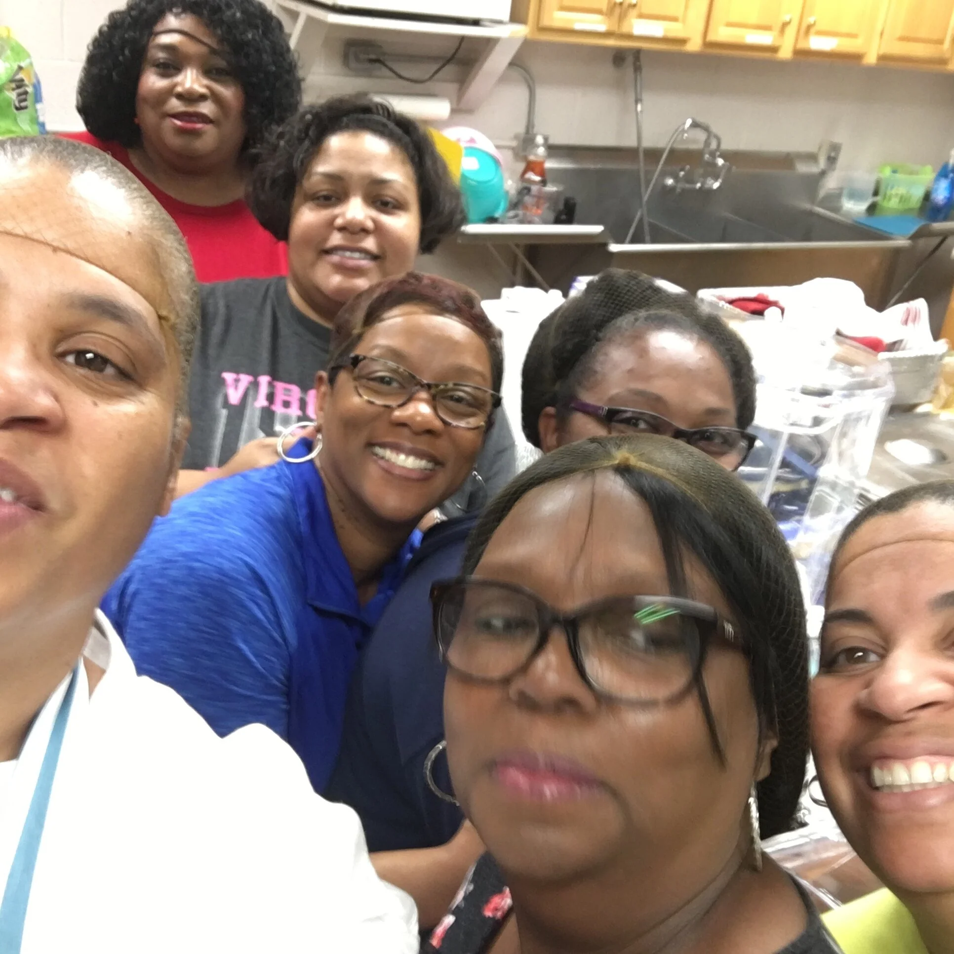 Group of women taking a selfie in a kitchen or food prep area, smiling and posing.