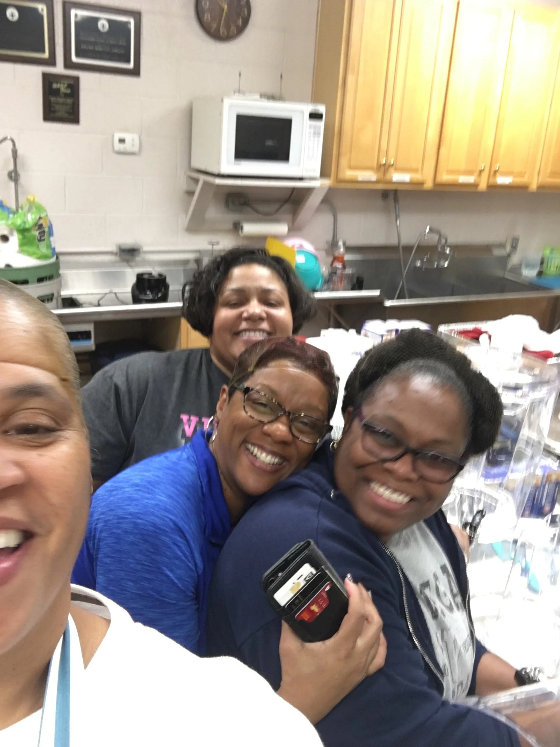 Four women smiling and taking a selfie in a kitchen with wooden cabinets, a microwave, and various kitchen items in the background.