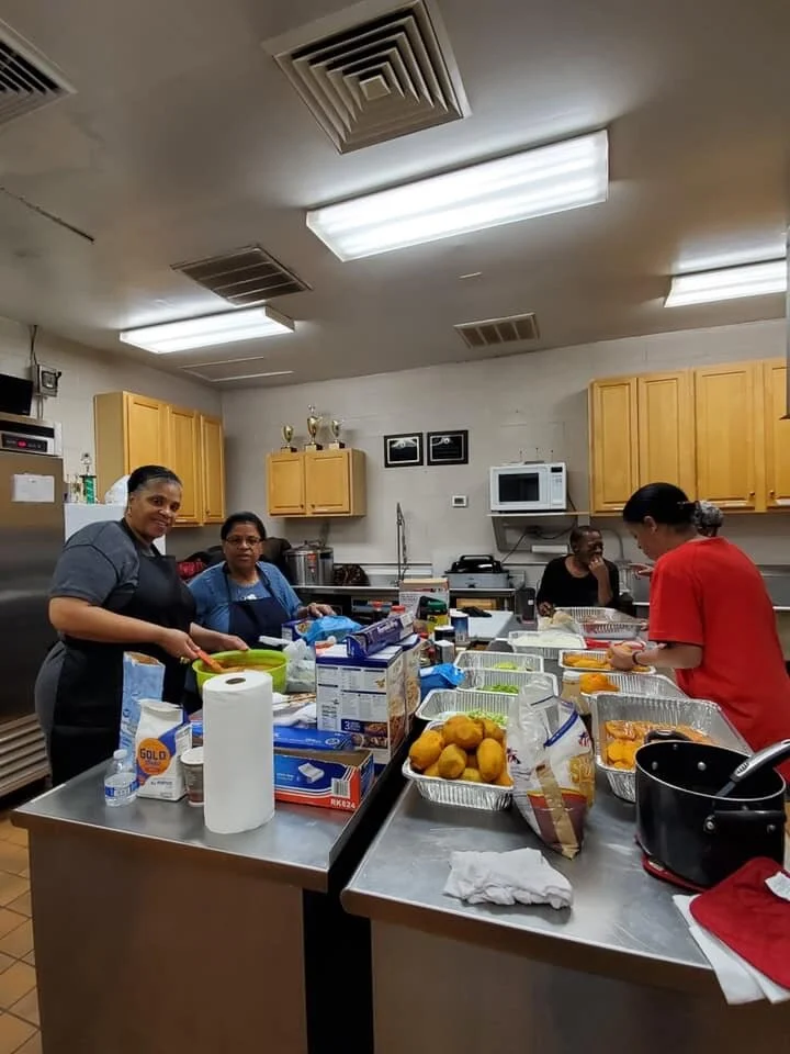 Group of women preparing food in a community kitchen with aluminum trays, lemons, and kitchen utensils on the counter.