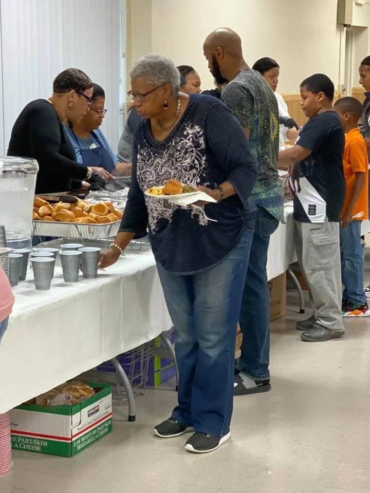 People lined up for food at a buffet table with bread rolls and fried chicken.