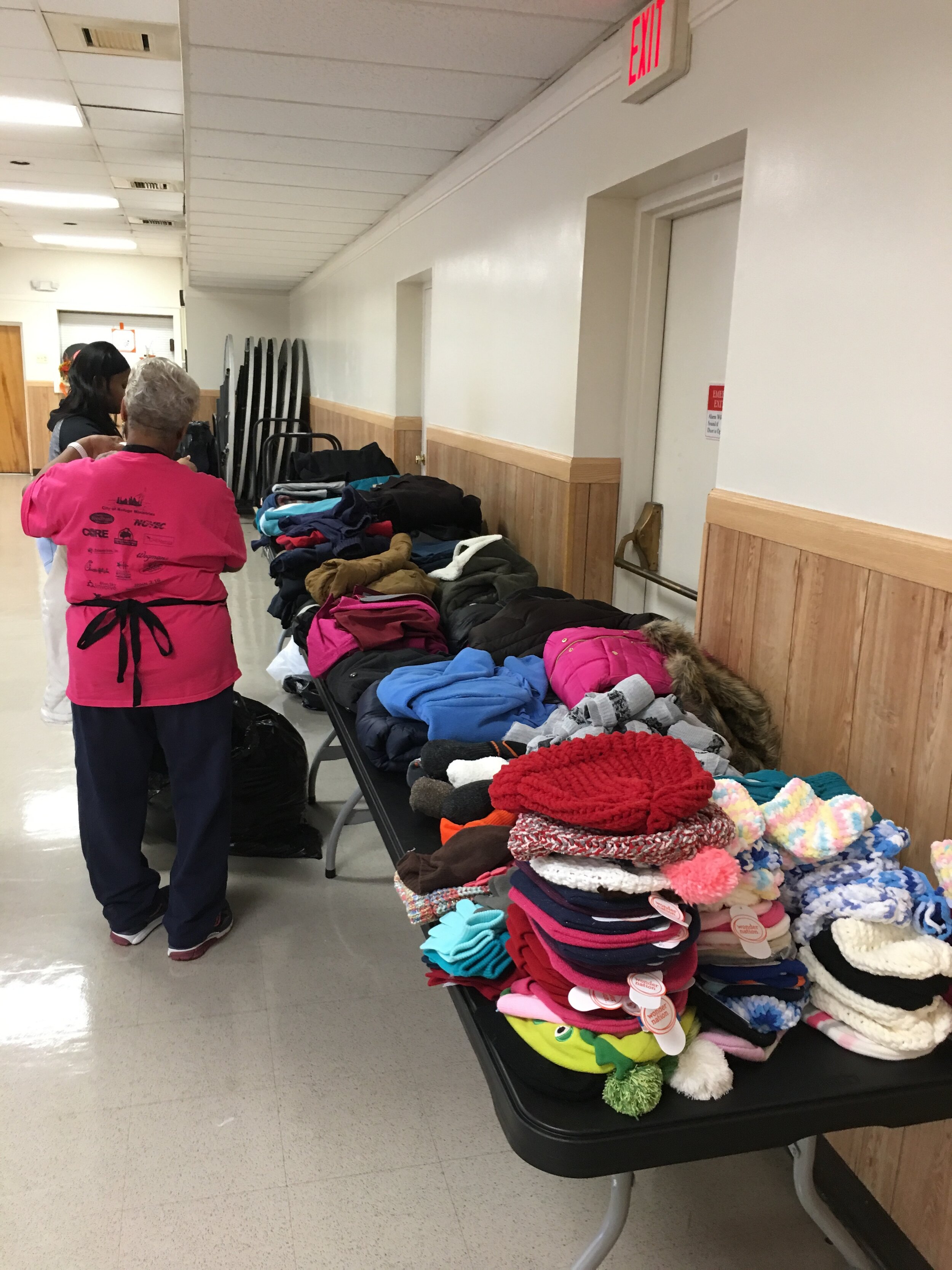 Table filled with various donated hats, gloves, and winter accessories, with two women organizing or selecting items.