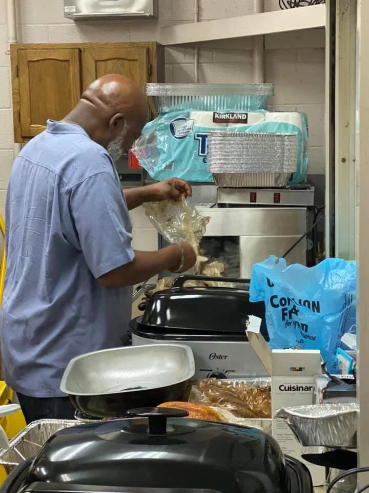 An older man with a bald head and gray beard wearing a blue shirt preparing food in a kitchen. He is standing in front of a small stove, handling what appears to be cooked chicken from a plastic bag. The kitchen has wooden cabinets, a white wall, and