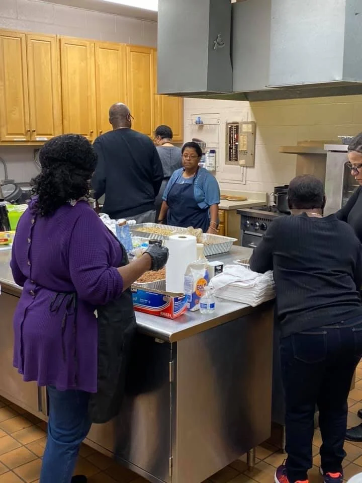 People working in a kitchen preparing food. Some are in aprons, and the kitchen has wooden cabinets and stainless steel appliances.